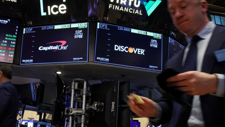 As telas exibem os logotipos e informações de negociação da Capital One Financial e da Discover Financial enquanto os traders trabalham no pregão da Bolsa de Valores de Nova York em 20 de fevereiro de 2024. (Foto: Brendan Mcdermid | Reuters)