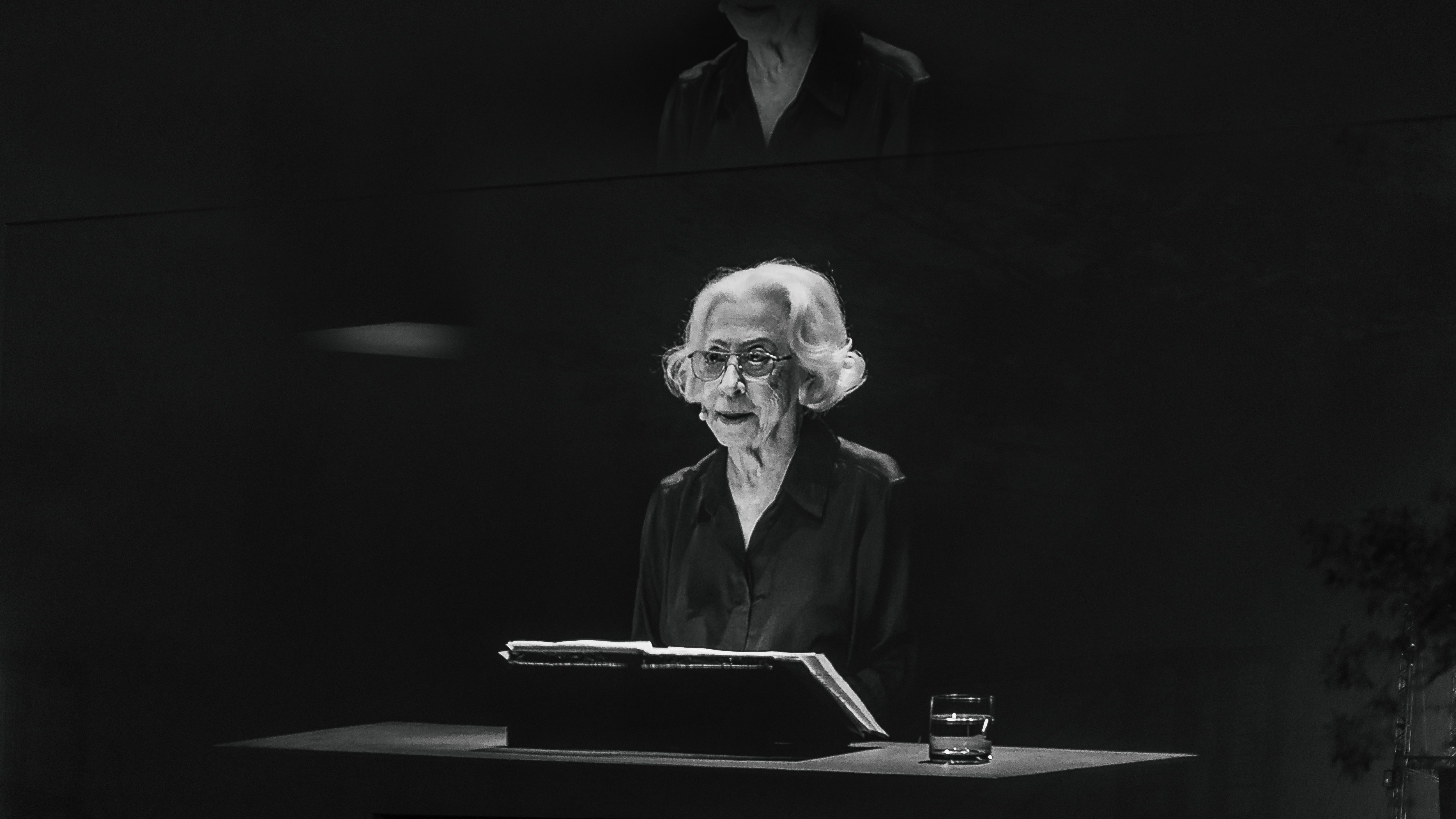 Foto preto e branca mostra Fernanda Montenegro, mulher idosa de cabelos brancos e lisos na altura do queixo. Ela usa óculos de grau e camisa preta. Está sentada diante de uma mesa preta. Há um livro aberto à sua frente, assim como um copo com água do seu lado direito. 