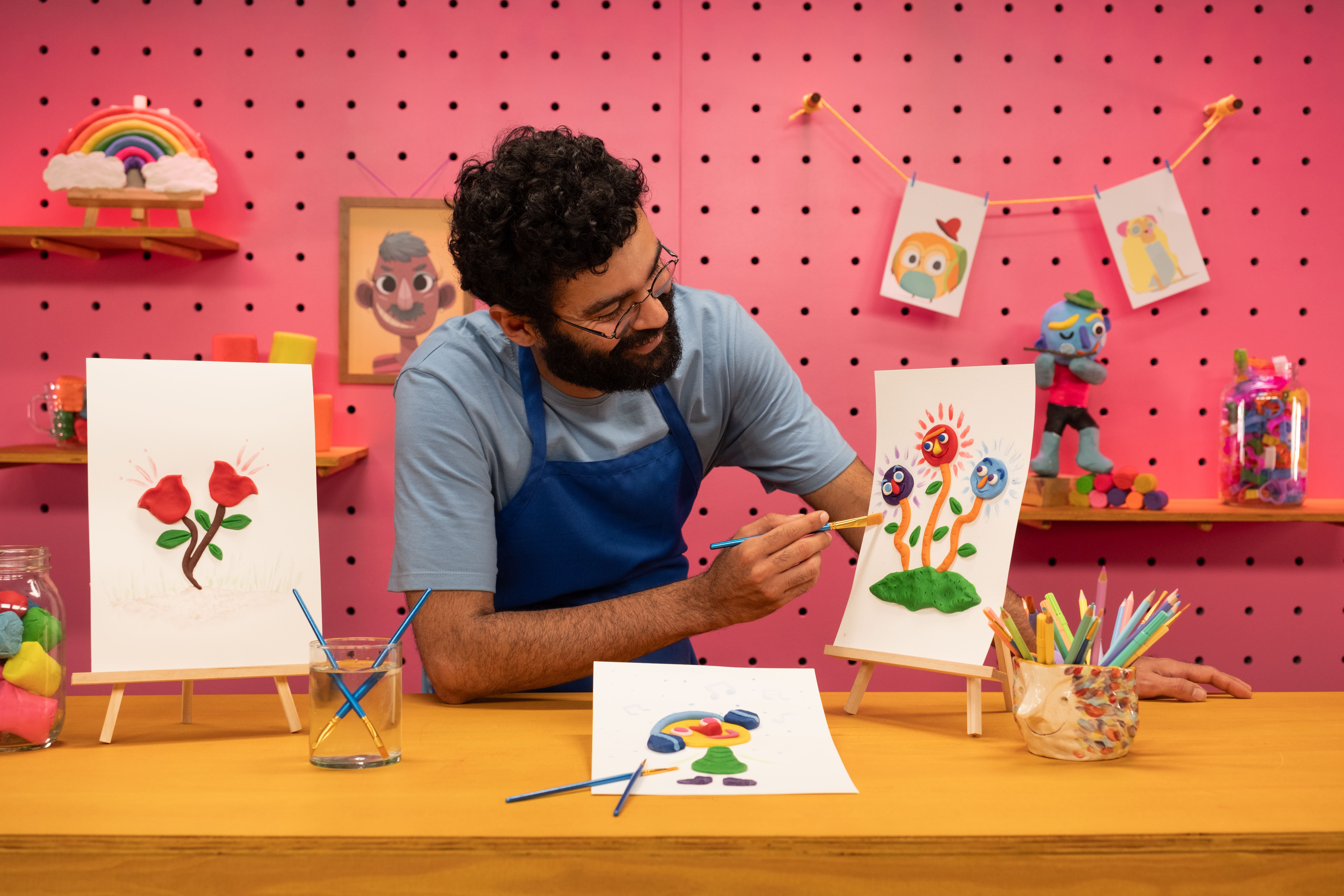 Na imagem um homem usa uma blusa azul e usa óculos, ele está segurando um pincel e um desenho.