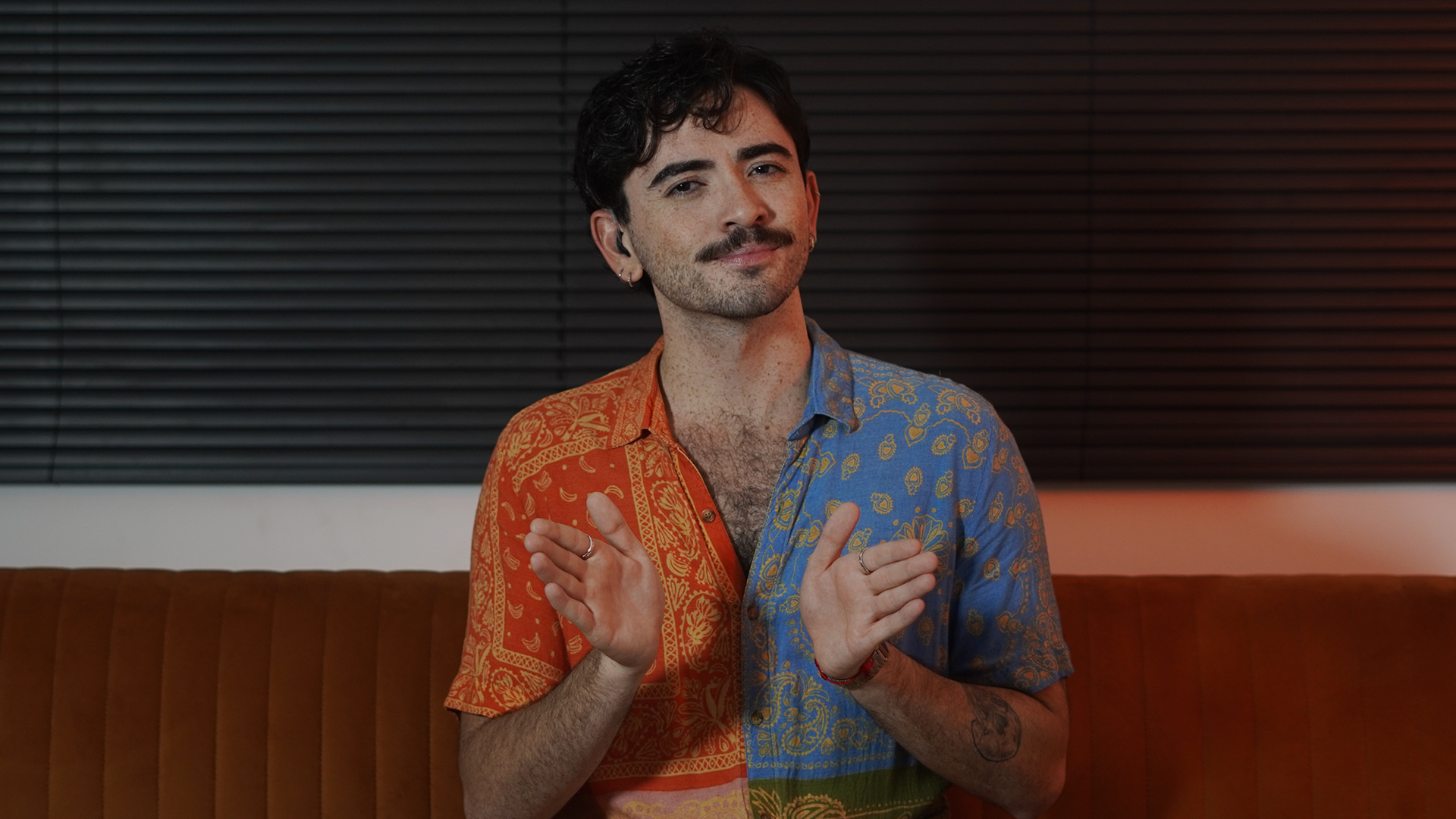 Homem jovem e branco aparece da cintura para cima, aparentemente sentado em um sofá de couro marrom. Ele tem cabelos curtos e pretos e usa bigode, também preto. Atrás dele, acima do sofá, tem uma persiana preta. Ele usa uma camisa de botão estampada e colorida. Tem uma tatuagem no braço e está sinalizando em Libras, gesticulando com as duas mãos. 