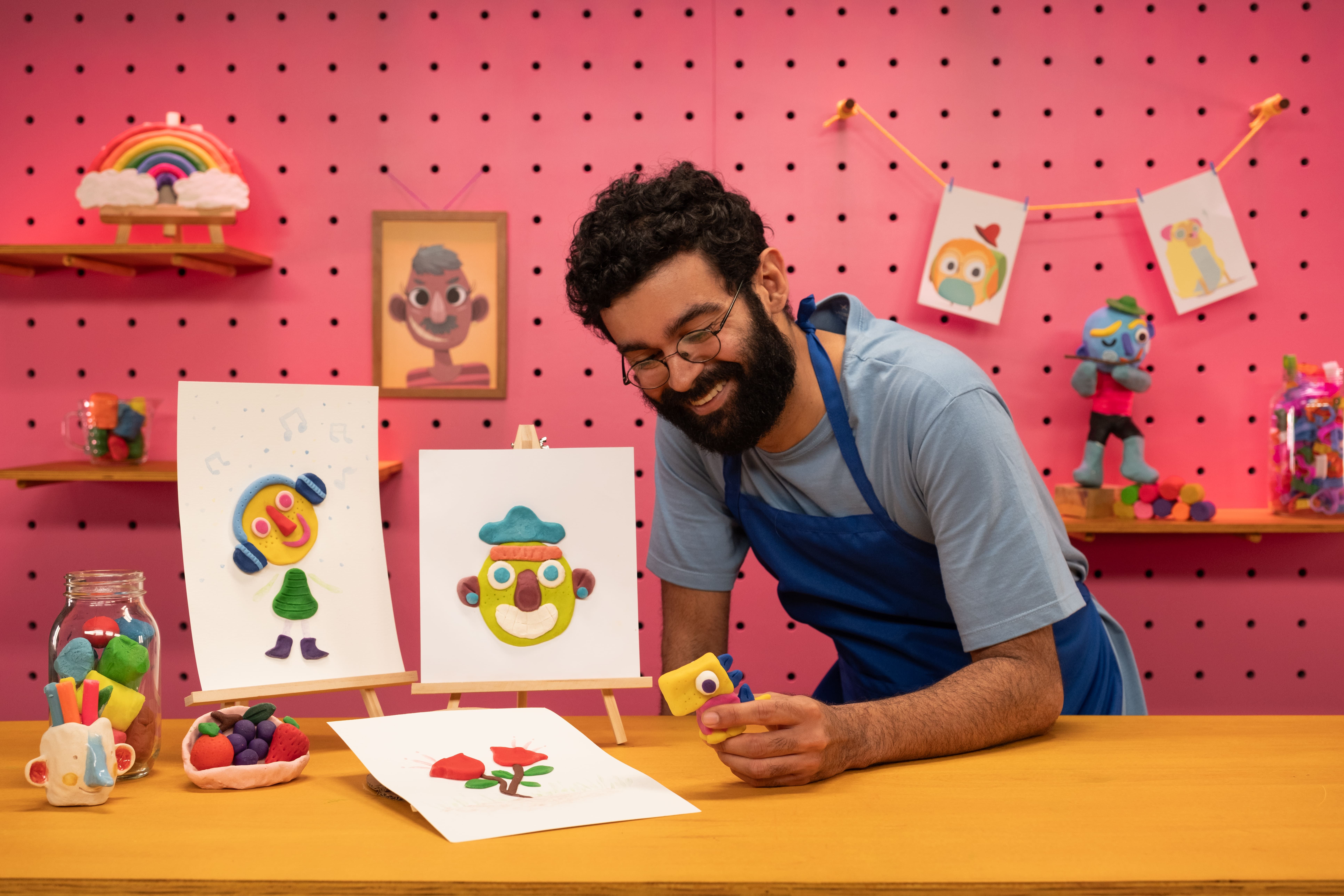 Na imagem um homem de cabelos crespos e preto usa óculos e tem barba, ele veste um avental azul e uma blusa de manga curta azul. Ele está com um dos braços em cima da mesa e segura na sua mão uma escultura de massinha.