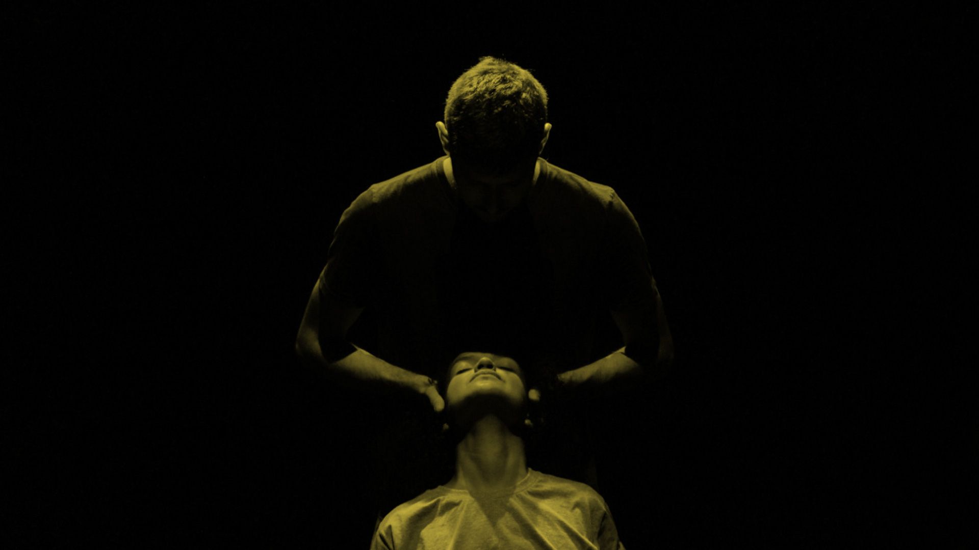 Um homem segura a cabeça de uma mulher. Ele está em pé, atrás dela. Ela está sentada. O fundo é preto.