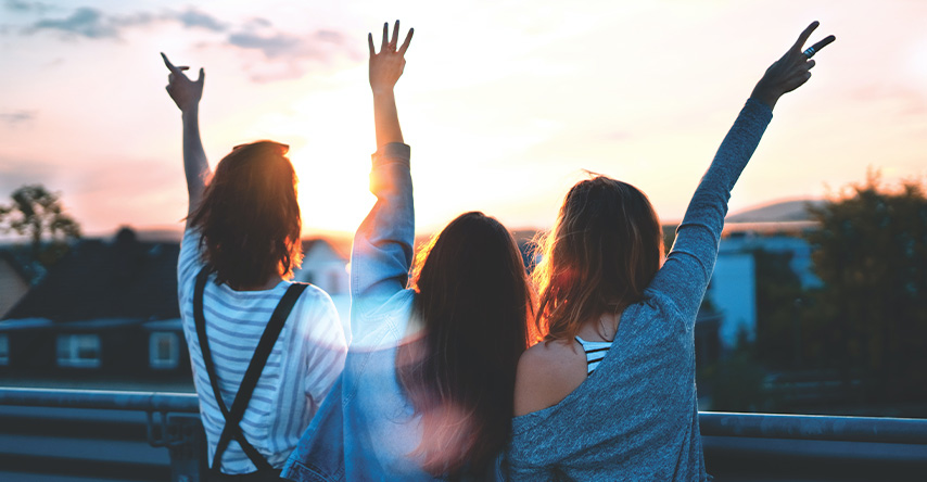 Grupo de três mulheres de costas olhando o nascer do sol de uma cidade.