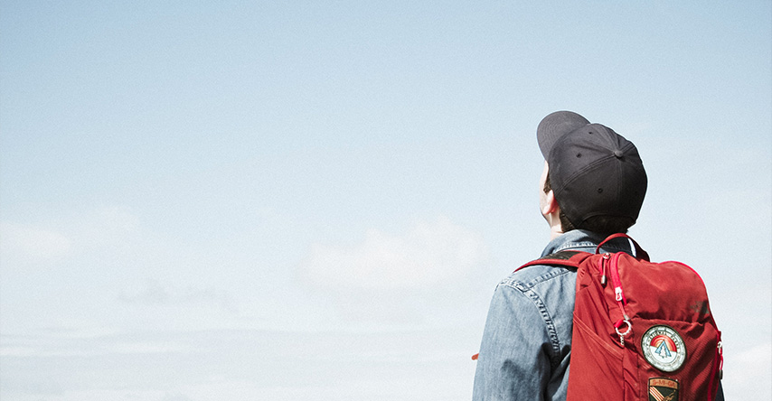 Homem de costas com um mochila vermelha olhando o céu.
