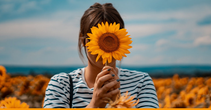 Mulher em um campo de girassóis segurando um girassol em frente ao seu rosto