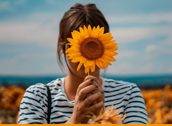 Mulher em um campo de girassóis segurando um girassol em frente ao seu rosto