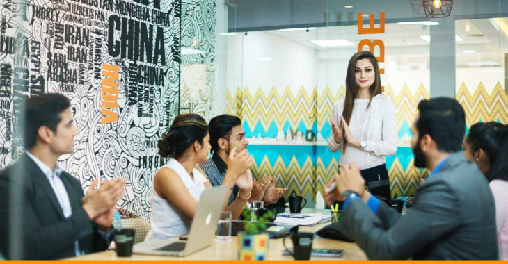 Mulher em pé e 5 pessoas sentadas ao redor de uma mesa batendo palmas após o término de uma reunião