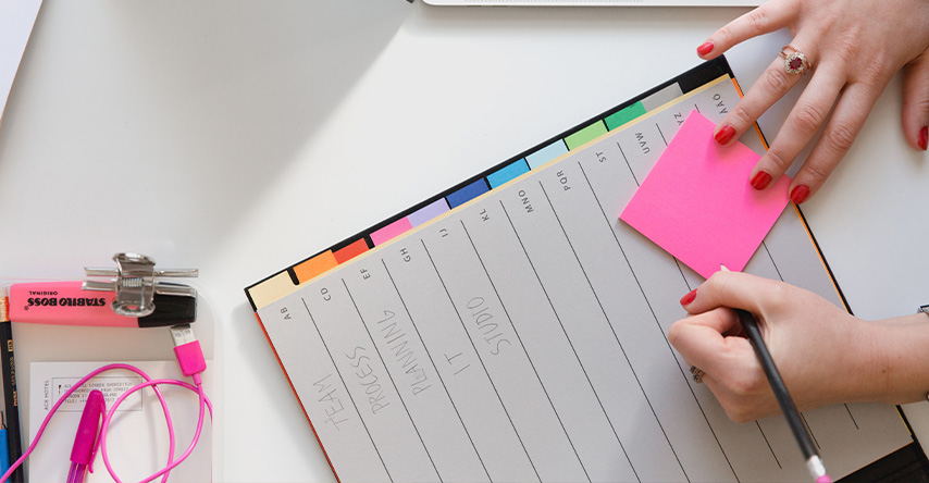 Moça escrevendo em um pos-it com um lápis em cima de uma agenda