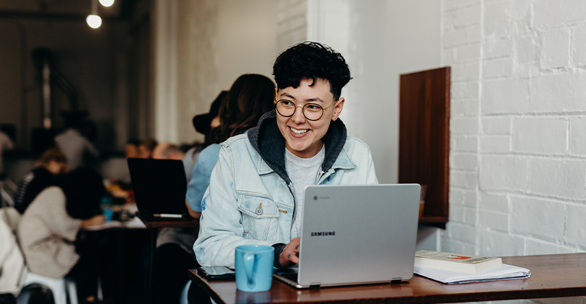Mulher sorrindo e olhando para o lado enquanto trabalha em seu computador em uma cafeteria.