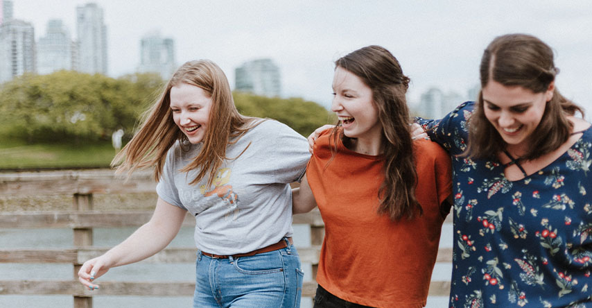 Três mulheres caminhando abraçadas lado a lado enquanto sorriem e se divertem.