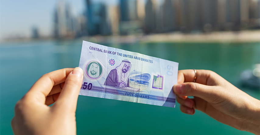 Mãos segurando nota de 50 dirhams, ao fundo a cidade de Dubai