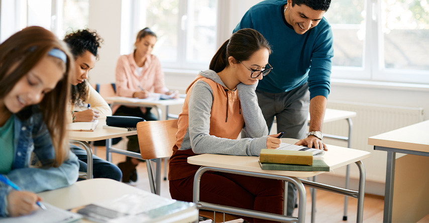 Adolescentes estudando em uma sala de aula enquanto o professor ajuda uma aluna.