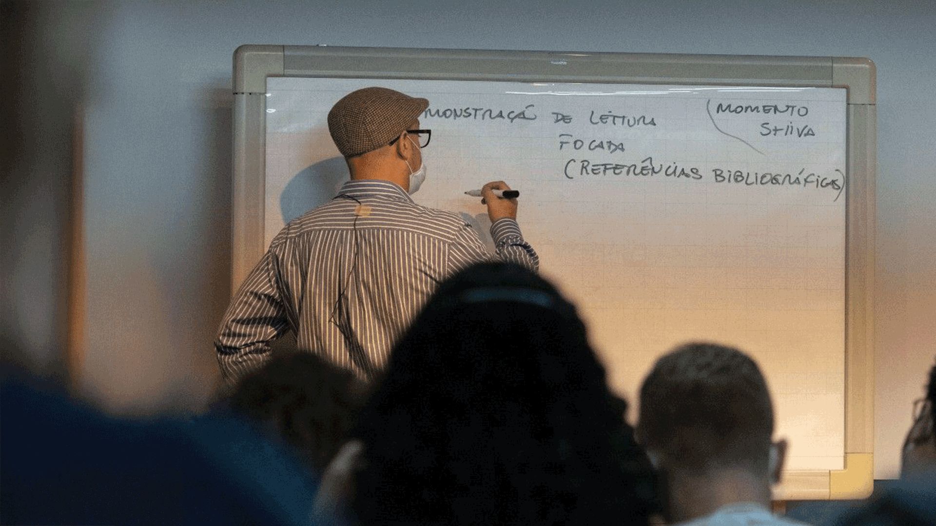 A fotografia é tirada como que do fundo de uma sala de aula. Além da cabeça e do torso de alunos sentados, vemos o professor, de costas, escrevendo na lousa. Ela usa boina e uma camisa listrada, é um homem branco.