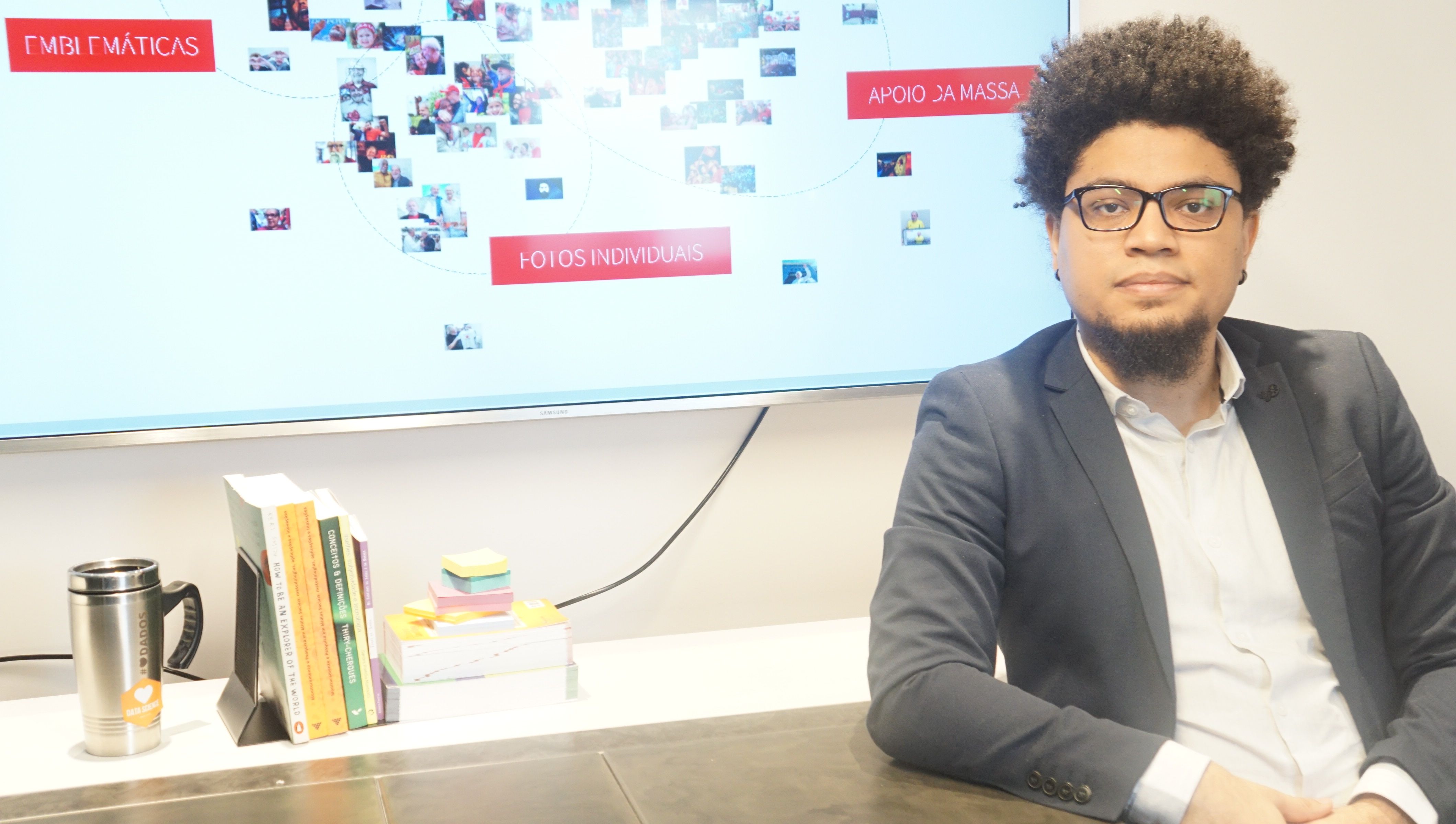 Na foto, o pesquisador Tarcízio Silva posa em frente à uma mesa com livros e ao fundo a projeção de uma apresentação.