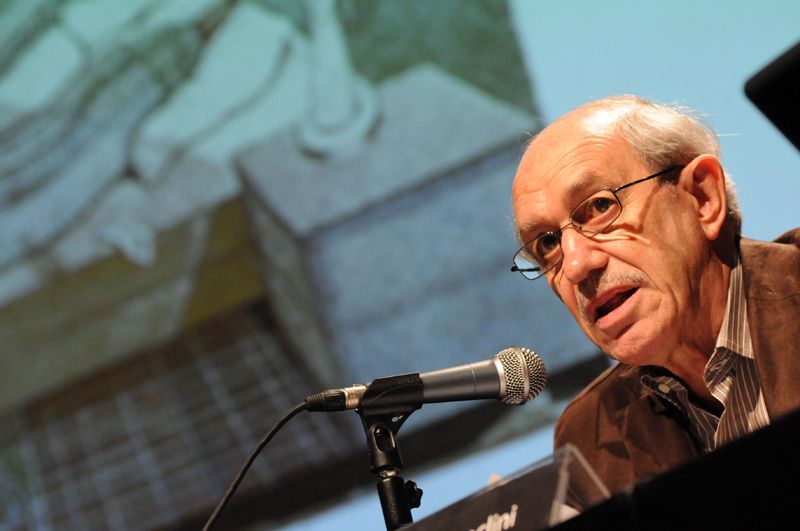 O antropólogo Néstor Garcia Canclini participa de um seminário. Ele está falando com o público, que não aparece na imagem. Canclini é um homem branco, calvo, de cabelos brancos, óculos e sem barba, com cerca de 80 anos.