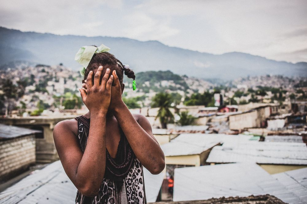 Uma mulher negra está com as mãos cobrindo os rostos. Ela veste uma blusa sem mangas e tem laços no cabelo. Ao fundo, de forma desfocada, há telhados e casas.