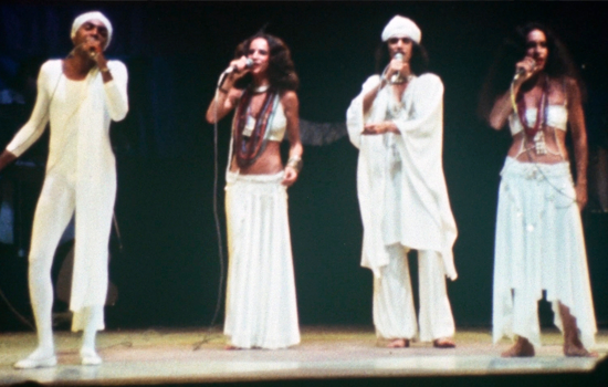 Na imagem est&atilde;o 4 pessoas, sendo dois homens e duas mulheres, Todos usam roupas brancas e cantam em um palco. 