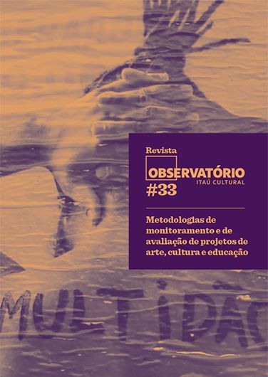 Ao fundo de uma parede azul com algumas manchas em vermelho, tem um cartaz de um homem, nú, negro, de costas, com as mãos entrelaçadas atrás da cabeça, um pouco mais abaixo nas costas escrito MULTIDÃO.