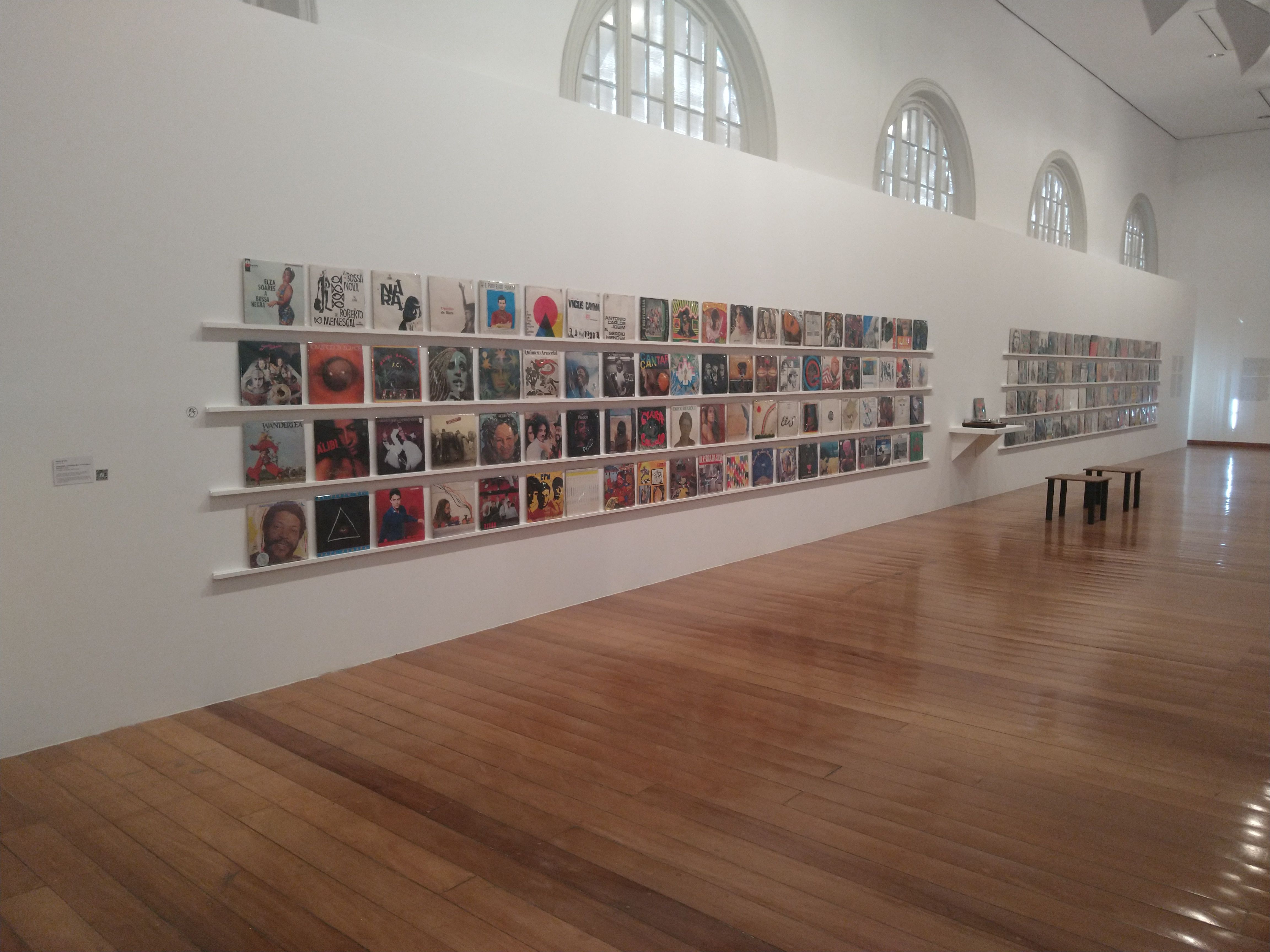 Fotografia colorida de uma parede com uma obra de arte exposta. A obra é composta por várias capas de discos de vinil organizados lado a lado em uma parede branca. Fotografia colorida de uma parede com uma obra de arte exposta. A obra é composta por várias capas de discos de vinil organizados lado a lado em uma parede branca.