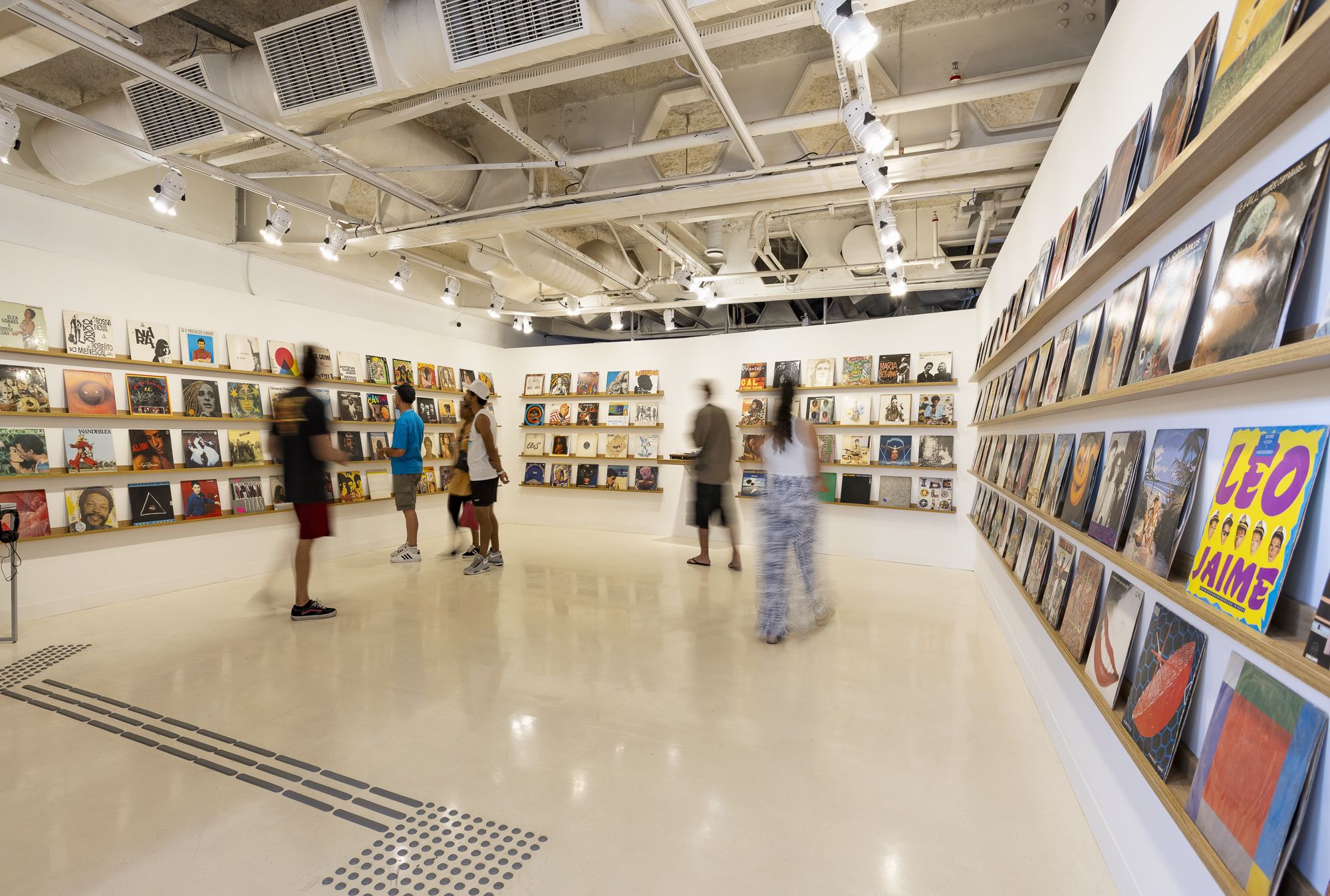 Fotografia de um espaço expositivo amplo e iluminado, com paredes brancas e teto industrial aparente, cheio de dutos e trilhos de luz. Nas paredes, prateleiras contínuas exibem dezenas de capas de discos de vinil, coloridas e organizadas lado a lado. No centro da sala, algumas pessoas caminham ou observam os discos; elas aparecem levemente borradas, sugerindo movimento. O piso é claro e liso, com sinalização tátil em relevo próxima ao primeiro plano.