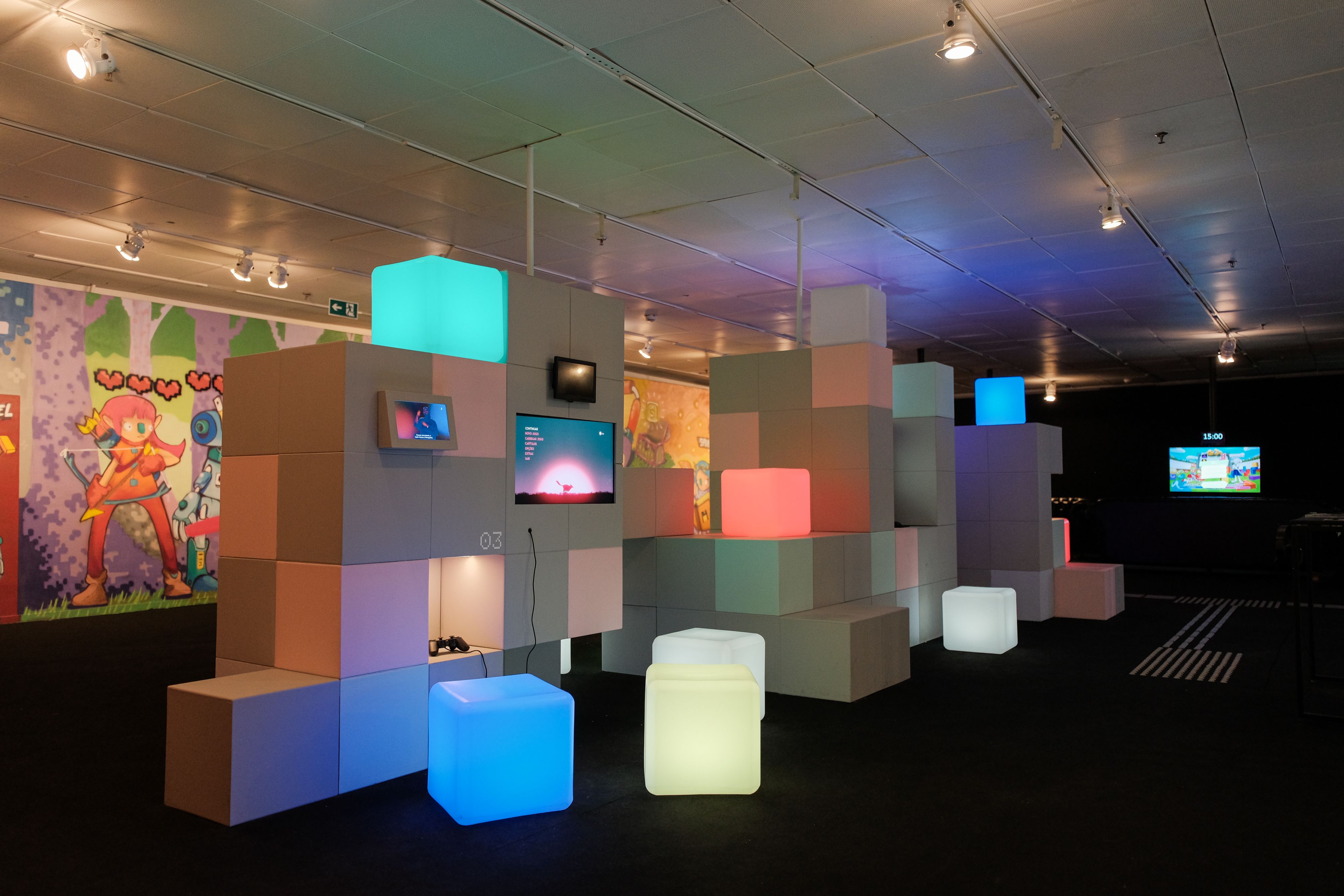 Imagem colorida mostra uma exposição interativa em ambiente interno, com iluminação baixa. No espaço, há várias estruturas feitas de grandes blocos cúbicos empilhados, em tons de cinza, bege e rosa. Alguns cubos emitem luz colorida — azul, verde, rosa e branco — funcionando como assentos ou elementos cenográficos. Em certas estruturas há telas exibindo jogos digitais. Ao fundo, paredes decoradas com ilustrações de personagens de videogame e, mais distante, um monitor contém um cronômetro indicando 15 minutos. O piso é escuro e o teto possui refletores alinhados. Imagem colorida mostra uma exposição interativa em ambiente interno, com iluminação baixa. No espaço, há várias estruturas feitas de grandes blocos cúbicos empilhados, em tons de cinza, bege e rosa. Alguns cubos emitem luz colorida — azul, verde, rosa e branco — funcionando como assentos ou elementos cenográficos. Em certas estruturas há telas exibindo jogos digitais. Ao fundo, paredes decoradas com ilustrações de personagens de videogame e, mais distante, um monitor contém um cronômetro indicando 15 minutos. O piso é escuro e o teto possui refletores alinhados.