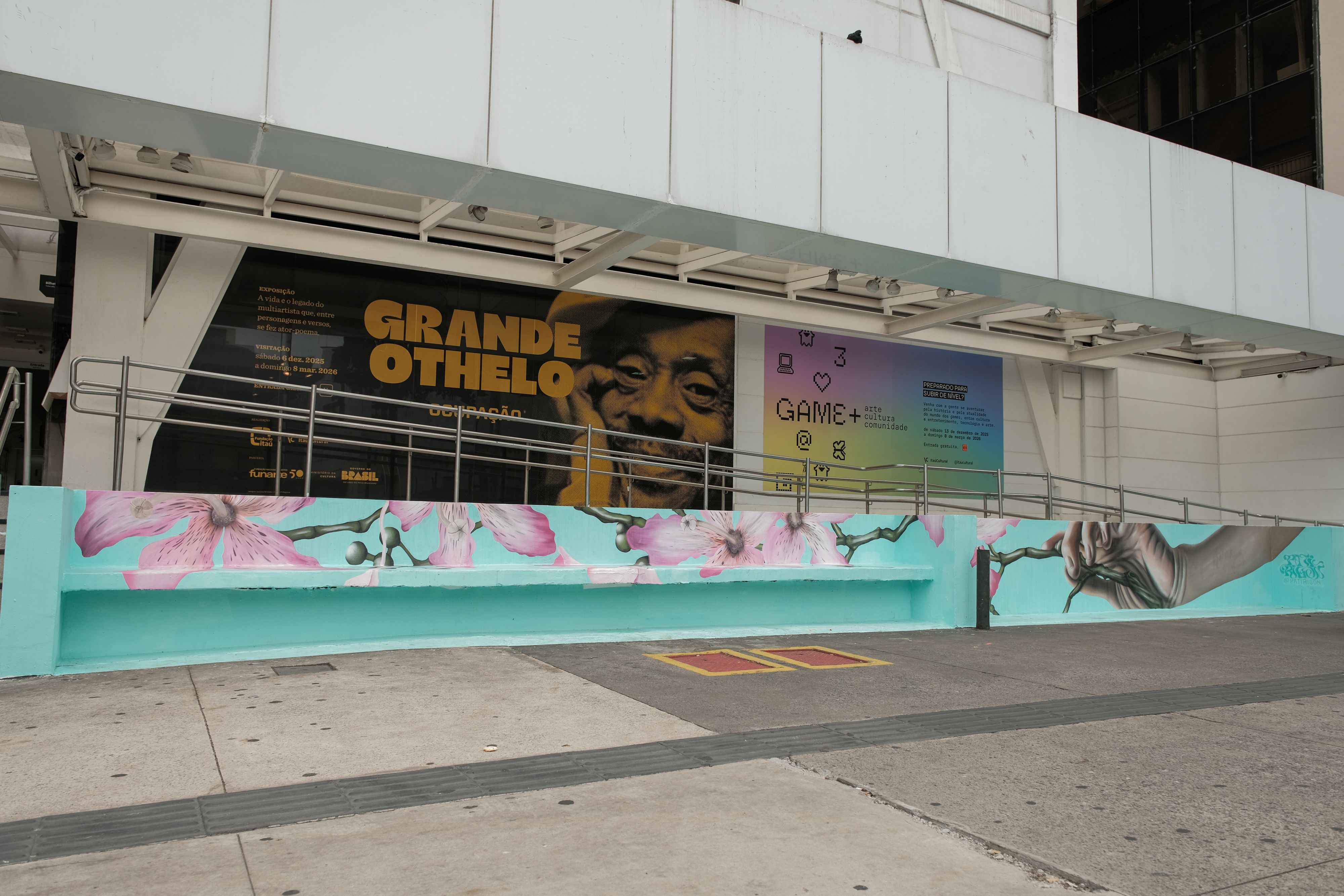 Fotografia da fachada do Itaú Cultural com uma intervenção de arte urbana. Em primeiro plano, um banco de concreto e uma mureta lateral foram pintados com fundo azul-claro e flores de pétalas rosadas. No canto direito da mureta, uma mão realista segura um galho verde.