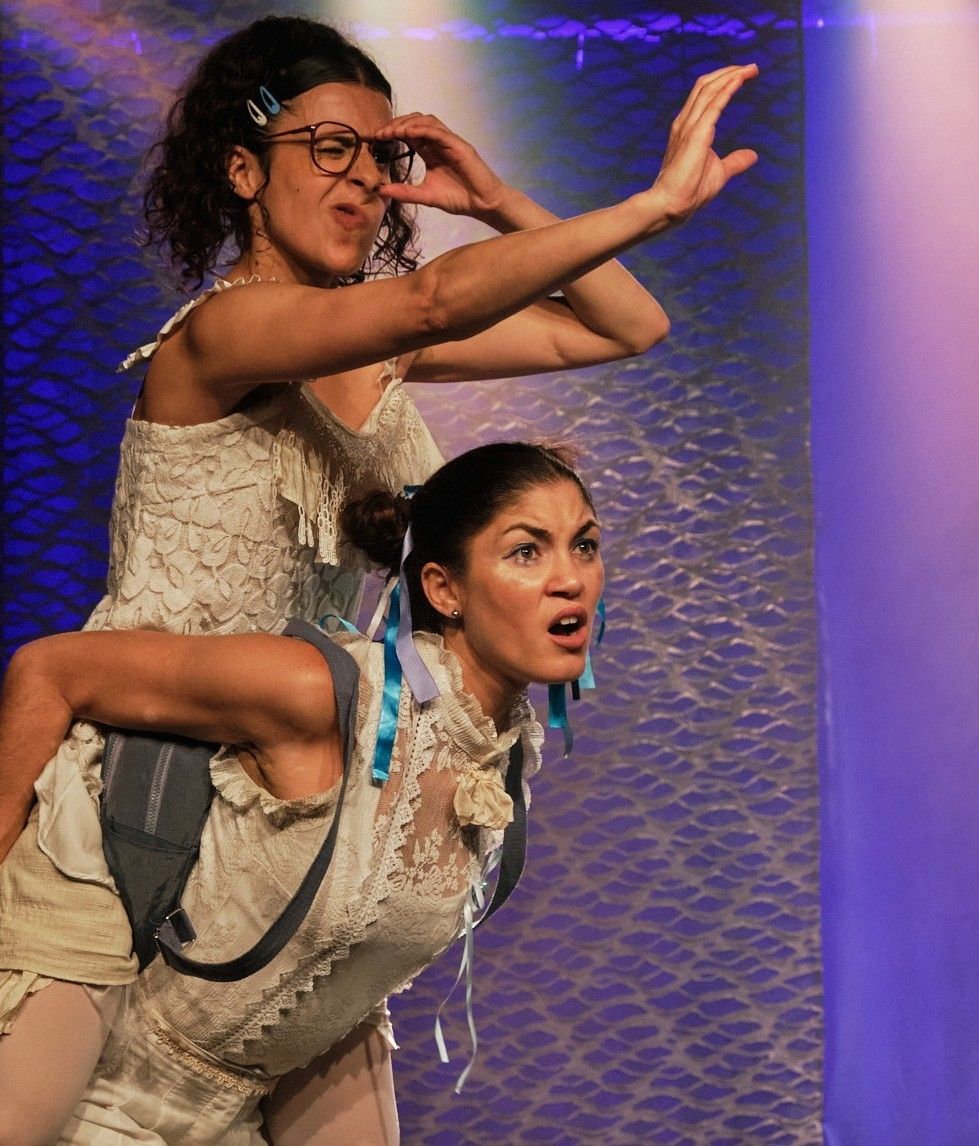 cena teatral vibrante e expressiva, onde duas atrizes vestidas com roupas de renda em tons de bege e branco protagonizam um momento de com&eacute;dia f&iacute;sica. A mulher &agrave; frente carrega a outra nas costas em um 