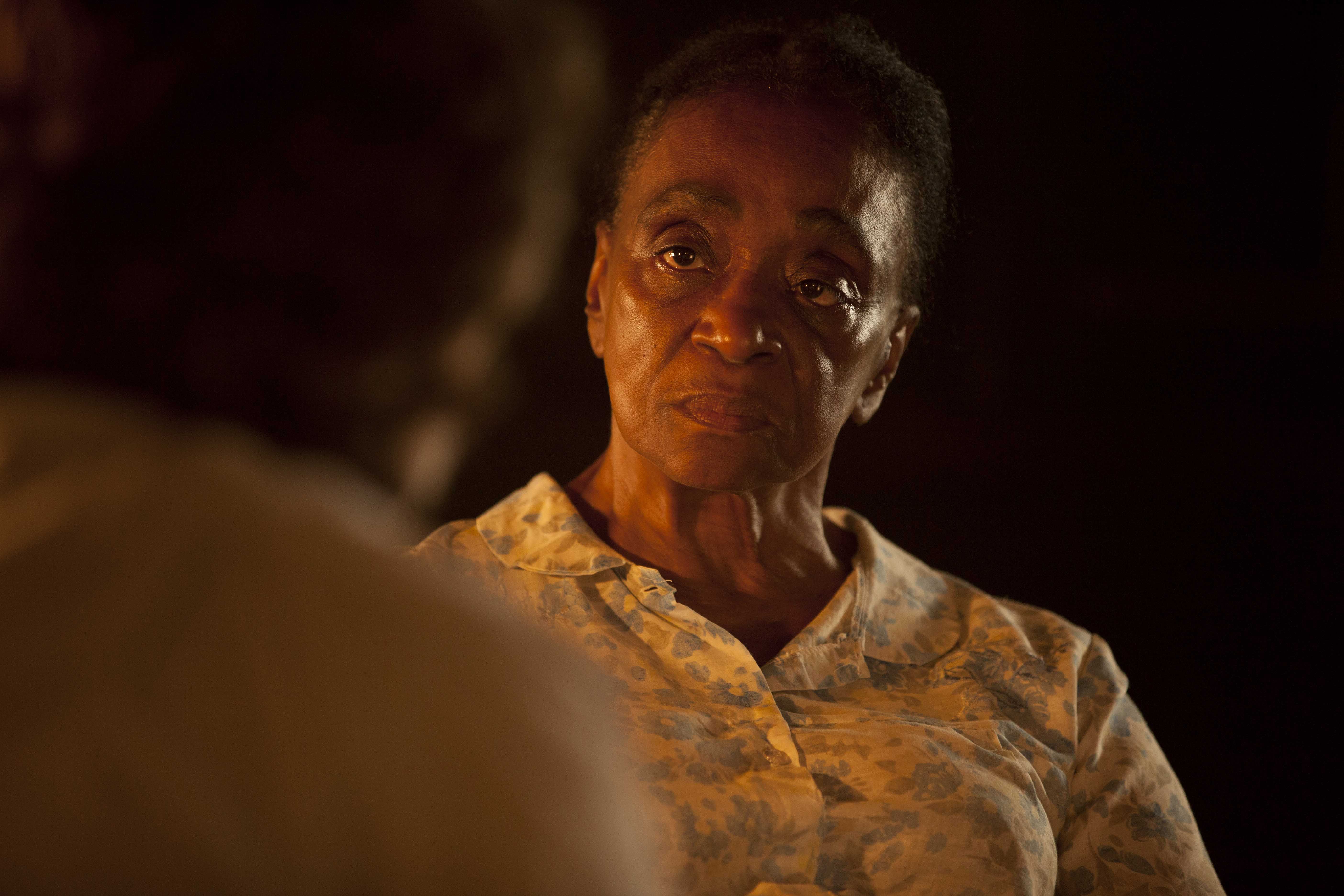 A imagem mostra a atriz L&eacute;a Garcia s&eacute;ria, olhando para outra pessoa. Ela &eacute; uma mulher negra e usa uma roupa branca com flores azuis.