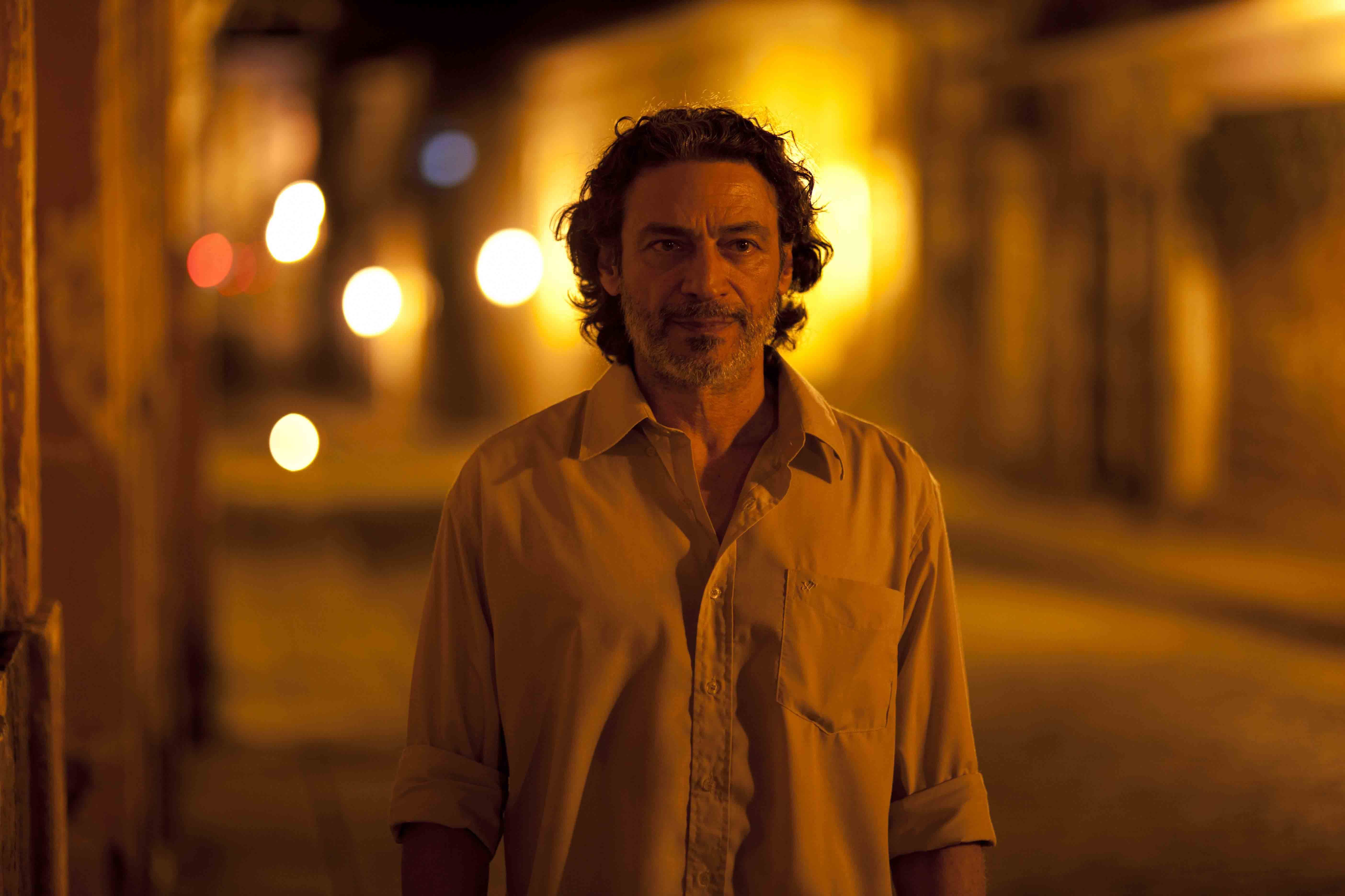 A imagem traz o ator Luiz Carlos Vasconcelos. Ele olha para frente, veste uma camisa, tem cabelo liso, barba e bigode. O artista &eacute; um homem branco. A foto apresenta uma luz amarelada.