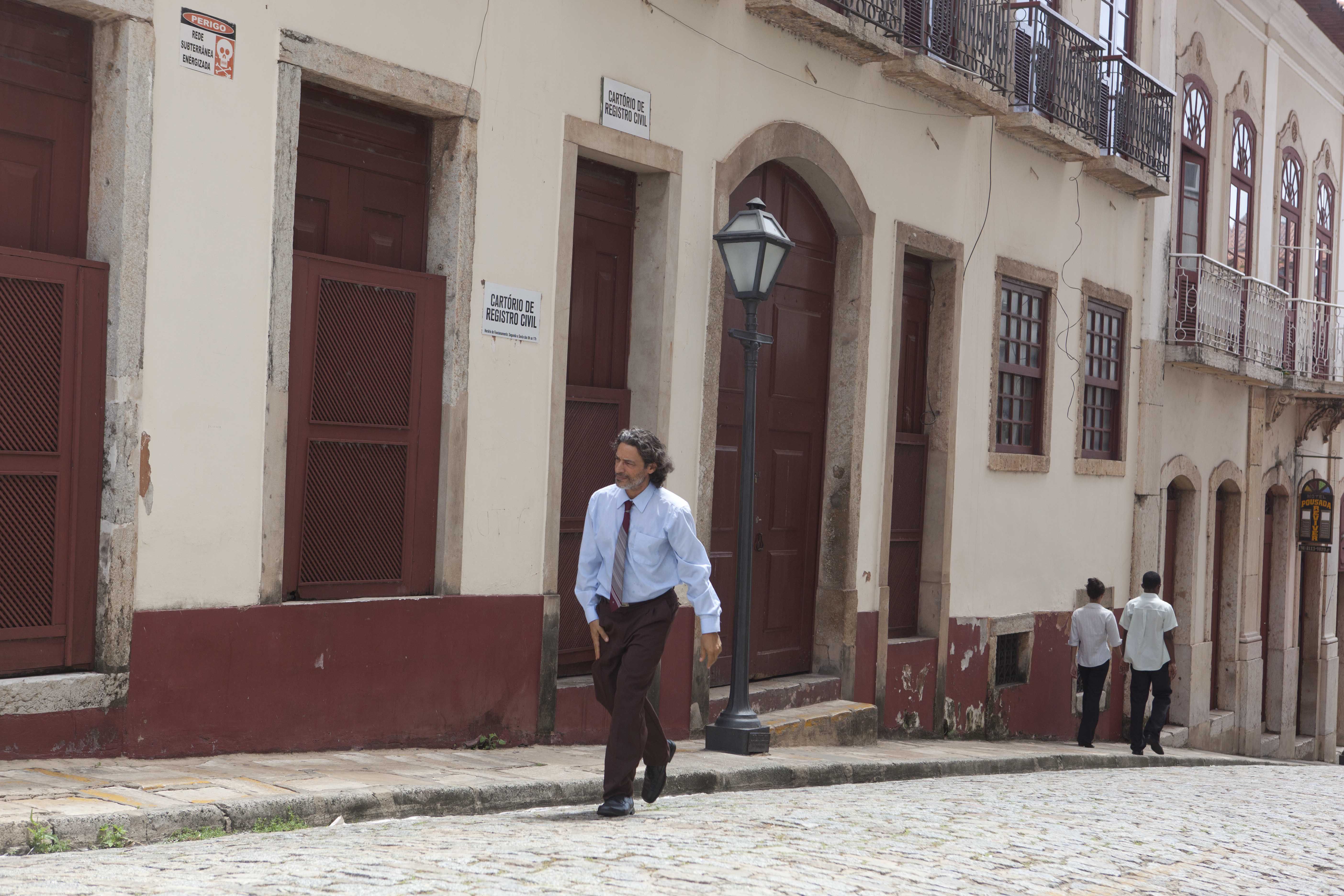 O ator Luiz Carlos Vasconcelos est&aacute; subindo uma ladeira de paralelep&iacute;pedos. Ele &eacute; um homem branco, veste uma camisa azul e uma cal&ccedil;a marrom. Ao fundo, h&aacute; um casar&atilde;o antigo e um poste de luz.