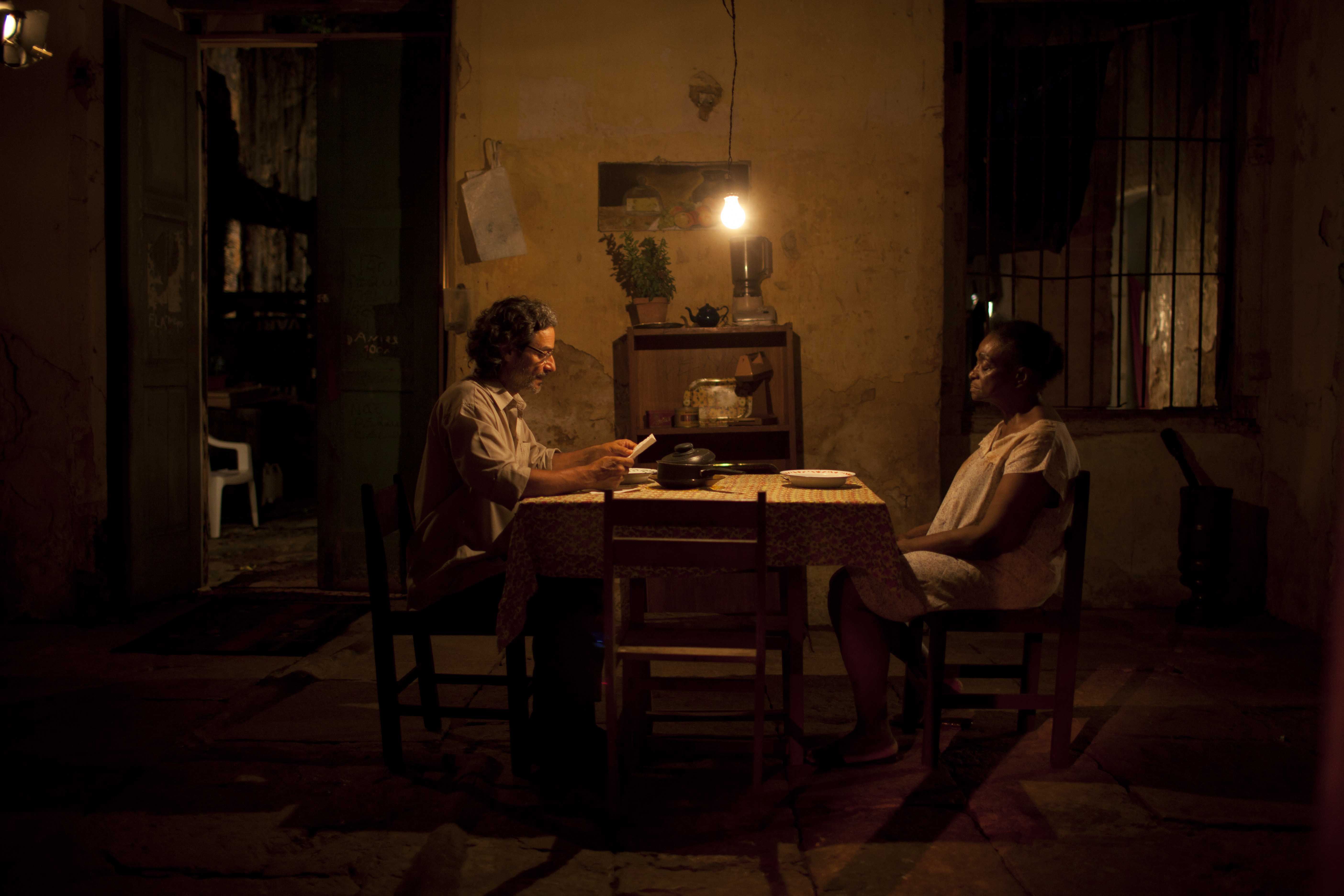 A imagem apresenta Léa Garcia e Luiz Carlos Vasconcelos em cena. Eles estão sentados à mesa. Ele, de óculos, lê algo para ela. Há uma lâmpada iluminando o ambiente doméstico.