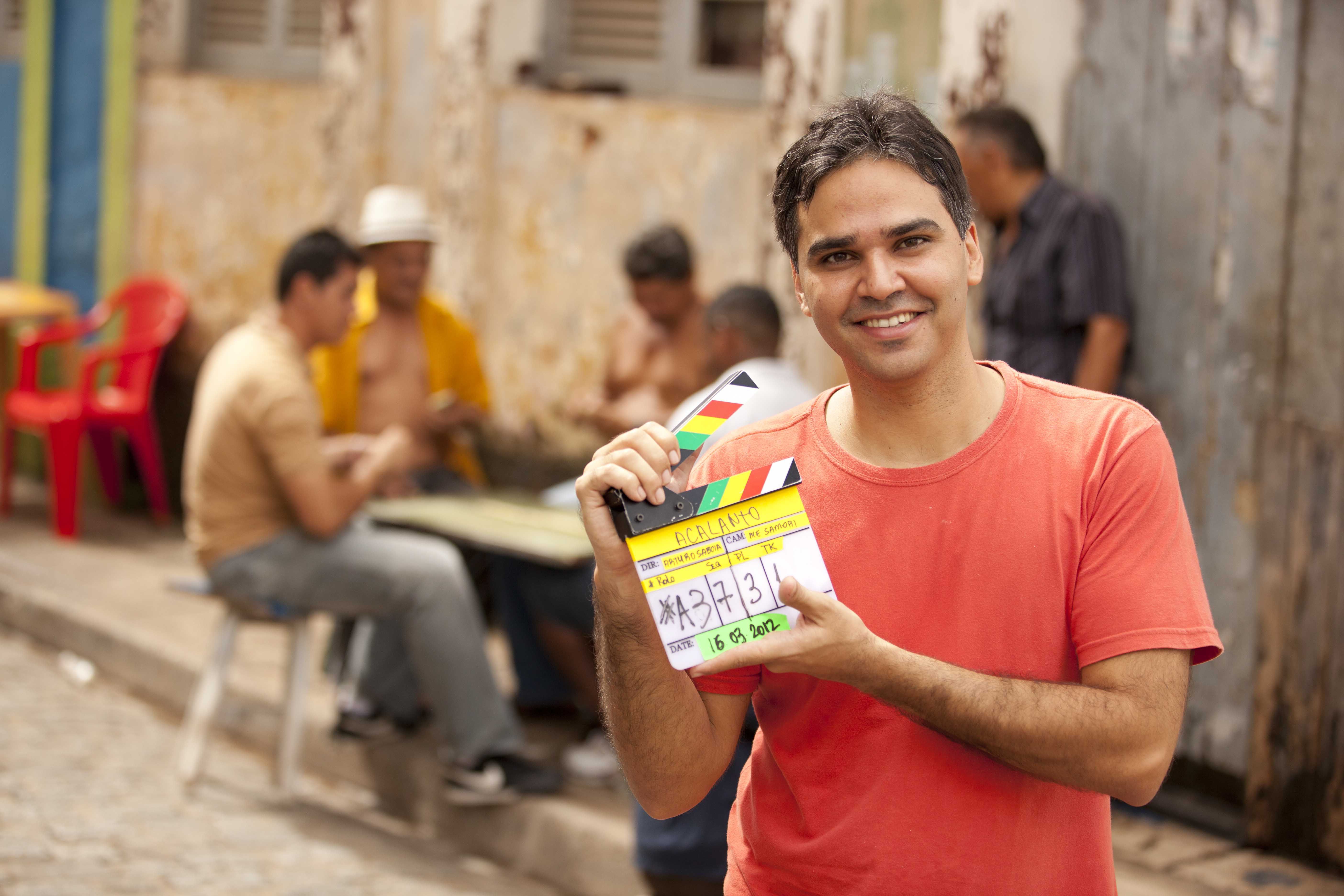 Arturo Saboia segura uma claquete em que aparece escrito o t&iacute;tulo do filme: &ldquo;Acalanto&rdquo;. O diretor veste uma camiseta vermelha. Arturo &eacute; branco, tem cabelo liso e escuro. Ao fundo, desfocados, homens est&atilde;o sentados em torno de uma mesinha, na rua.