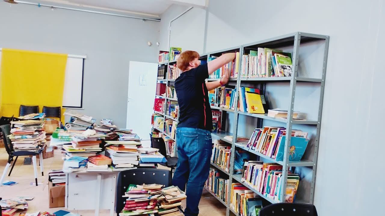 Imagem mostra o bibliotec&aacute;rio Osmar arrumando os livros na prateleira. Ao lado, pr&oacute;ximo dele, est&aacute; a mesa de trabalho com dezenas de livros em cima. 