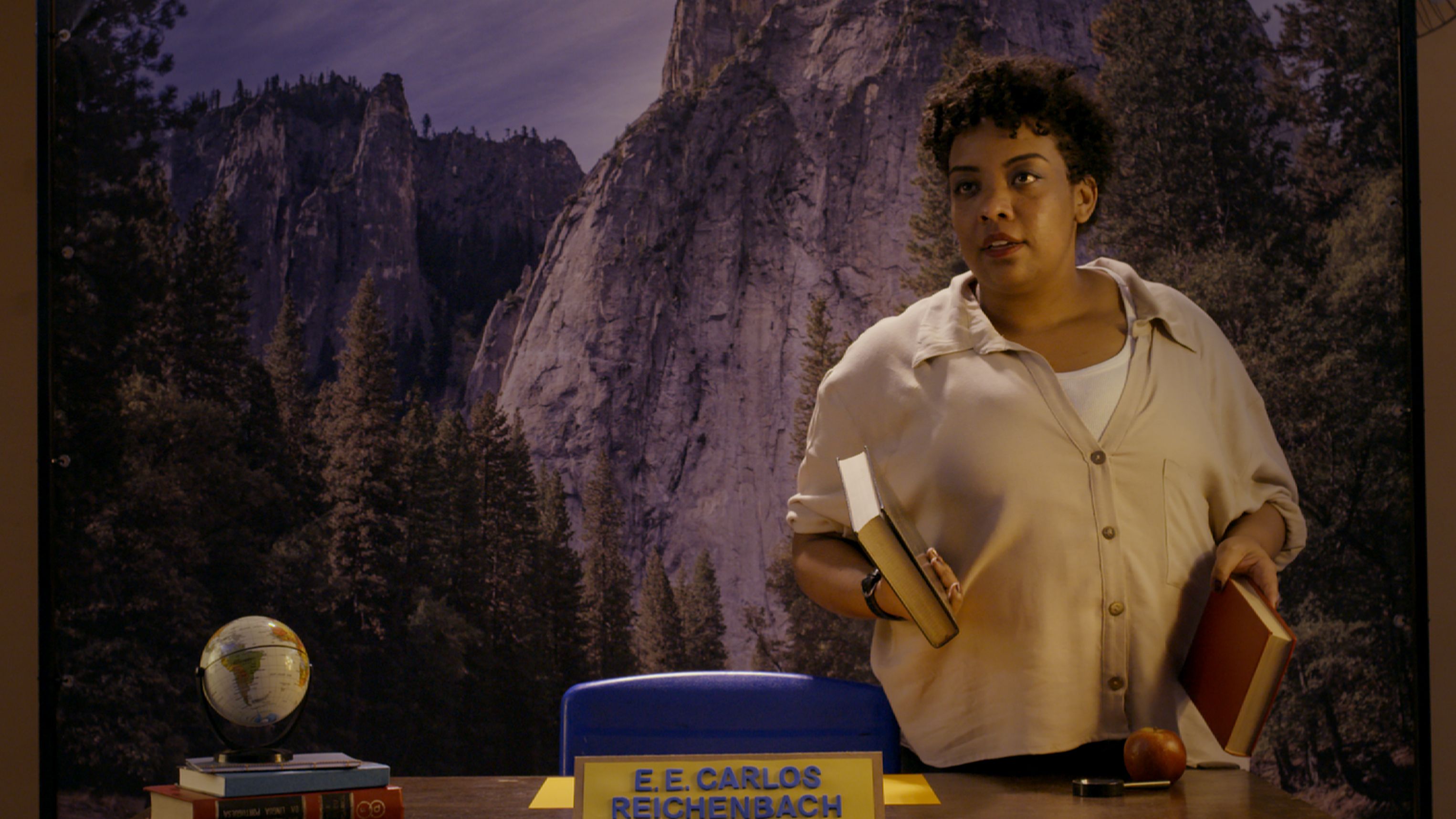 uma mulher de cabelos curtos e cacheados segurando dois livros enquanto est&aacute; em p&eacute; atr&aacute;s de uma mesa. Sobre a bancada, h&aacute; elementos que lembram um ambiente escolar, como um pequeno globo terrestre, uma pilha de livros, uma ma&ccedil;&atilde;, uma lupa e uma placa amarela com a inscri&ccedil;&atilde;o 