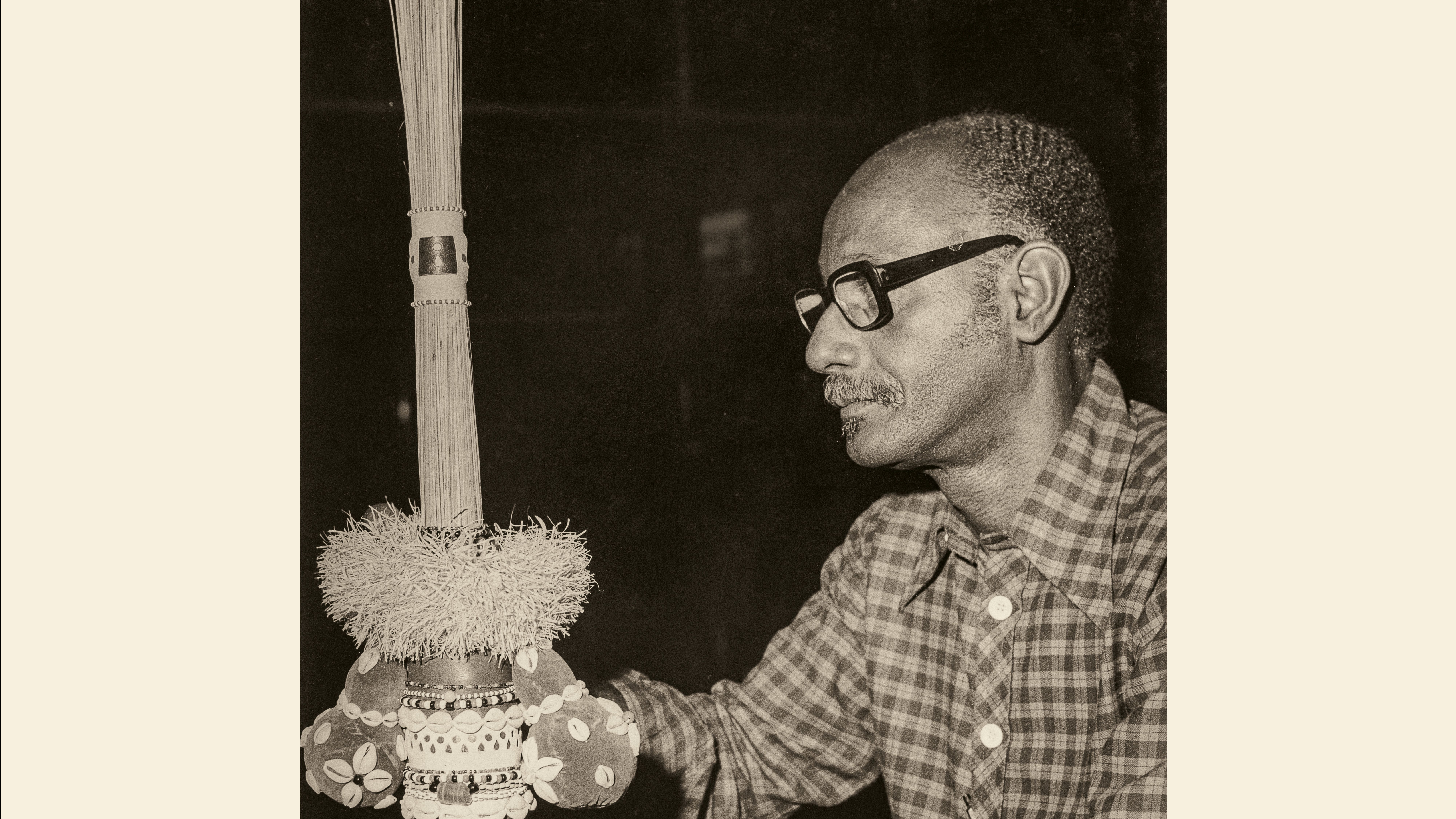 A foto em preto e branco traz Mestre Didi de perfil, usando óculos e uma camisa xadrez. Ele segura uma escultura de sua autoria.