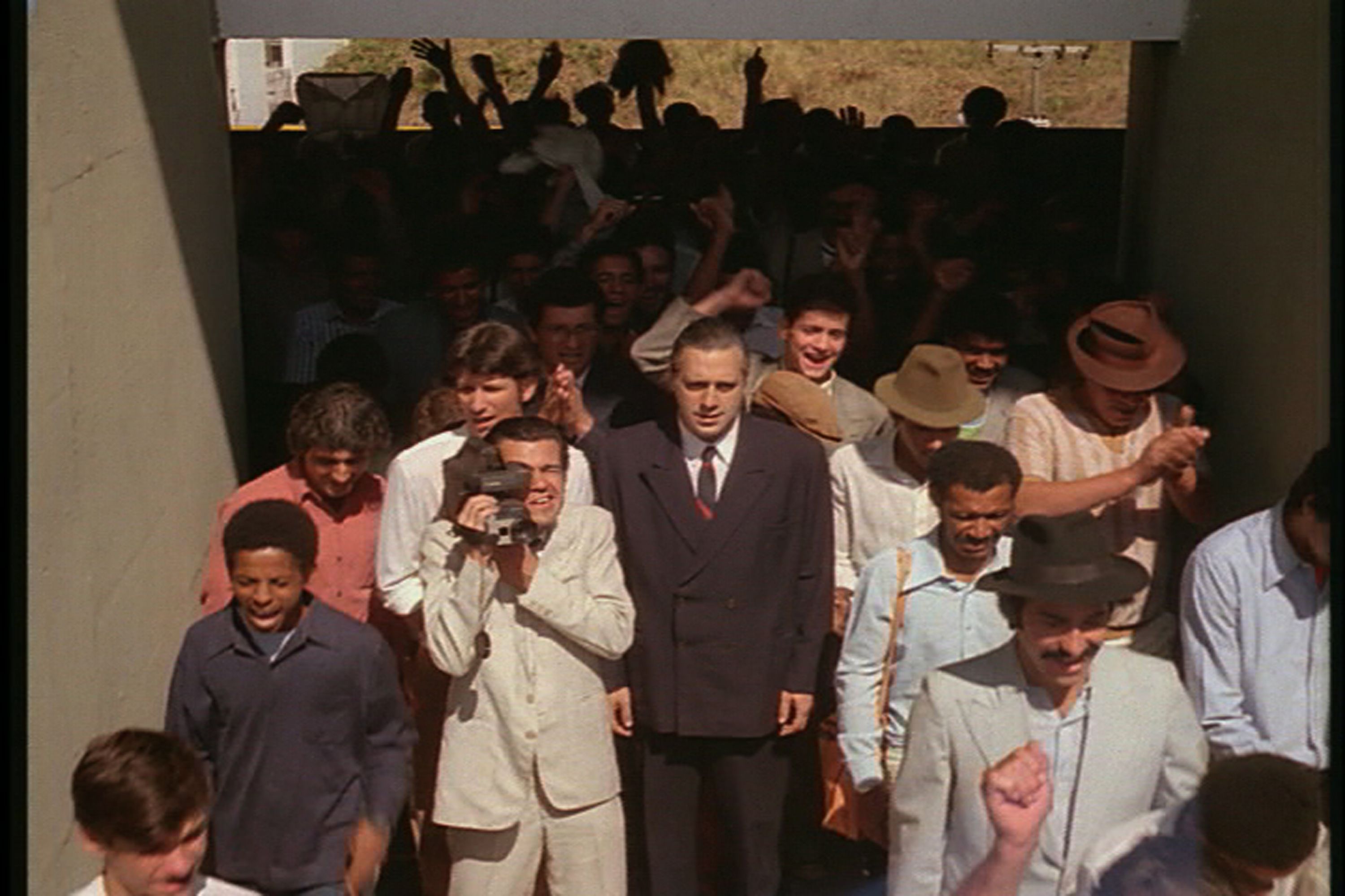 Ant&ocirc;nio Fagundes aparece de terno e gravata pretos e camisa branca. Ele est&aacute; s&eacute;rio e caminha com outros homens e garotos. Um deles segura uma gravadora.