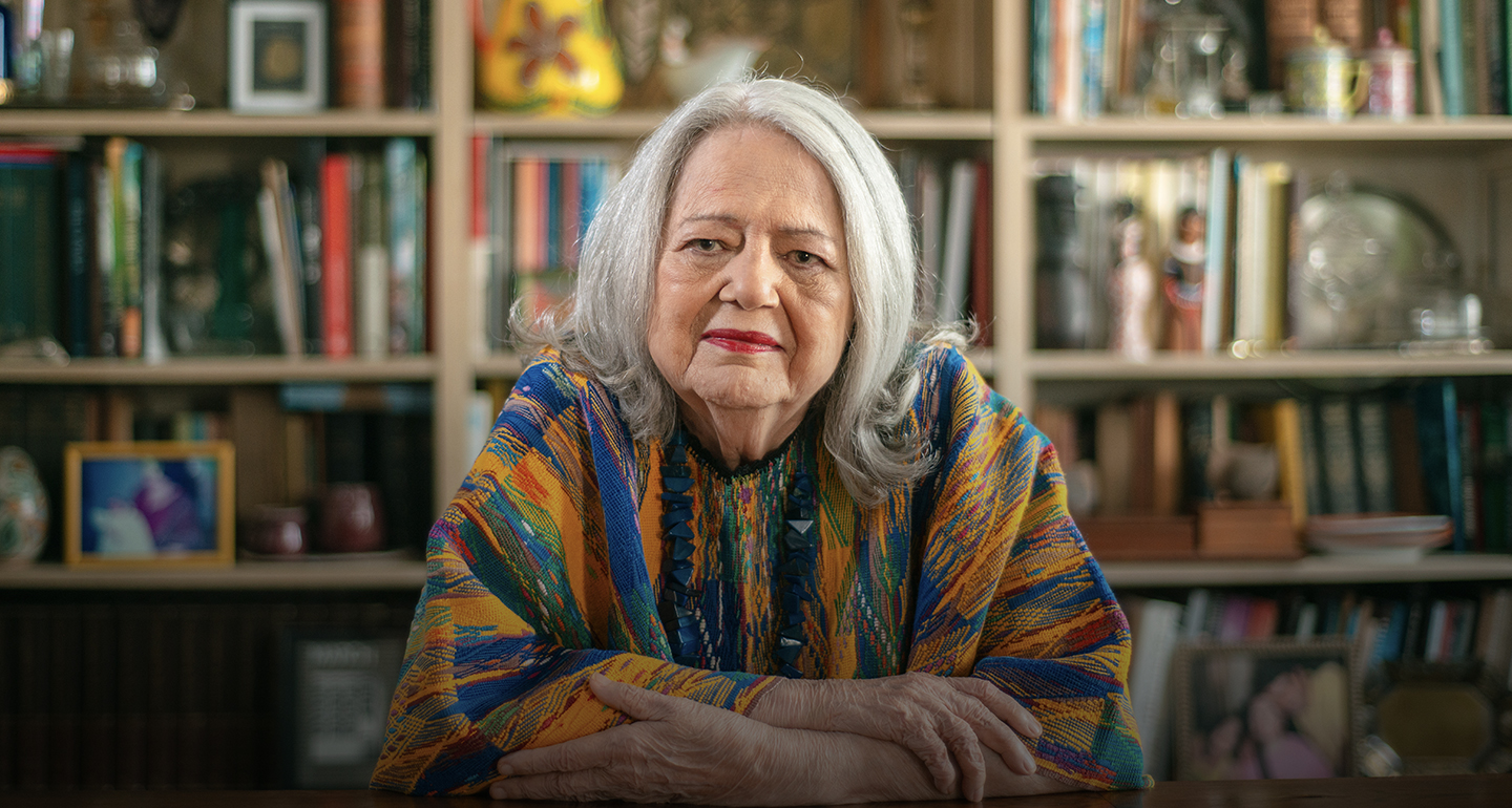 Fotografia em plano médio frontal da arte-educadora Ana Mae Barbosa. Uma mulher branca de cabelos longos, lisos e grisalhos, com expressão serena. Ela veste uma túnica colorida com padrões geométricos em tons de amarelo, azul, laranja e verde, e usa um colar de pedras azuis escuras. Seus braços estão cruzados sobre uma mesa de madeira em primeiro plano. Ao fundo, uma estante de livros repleta de obras e objetos decorativos está levemente desfocada, criando uma atmosfera intelectual e acolhedora.