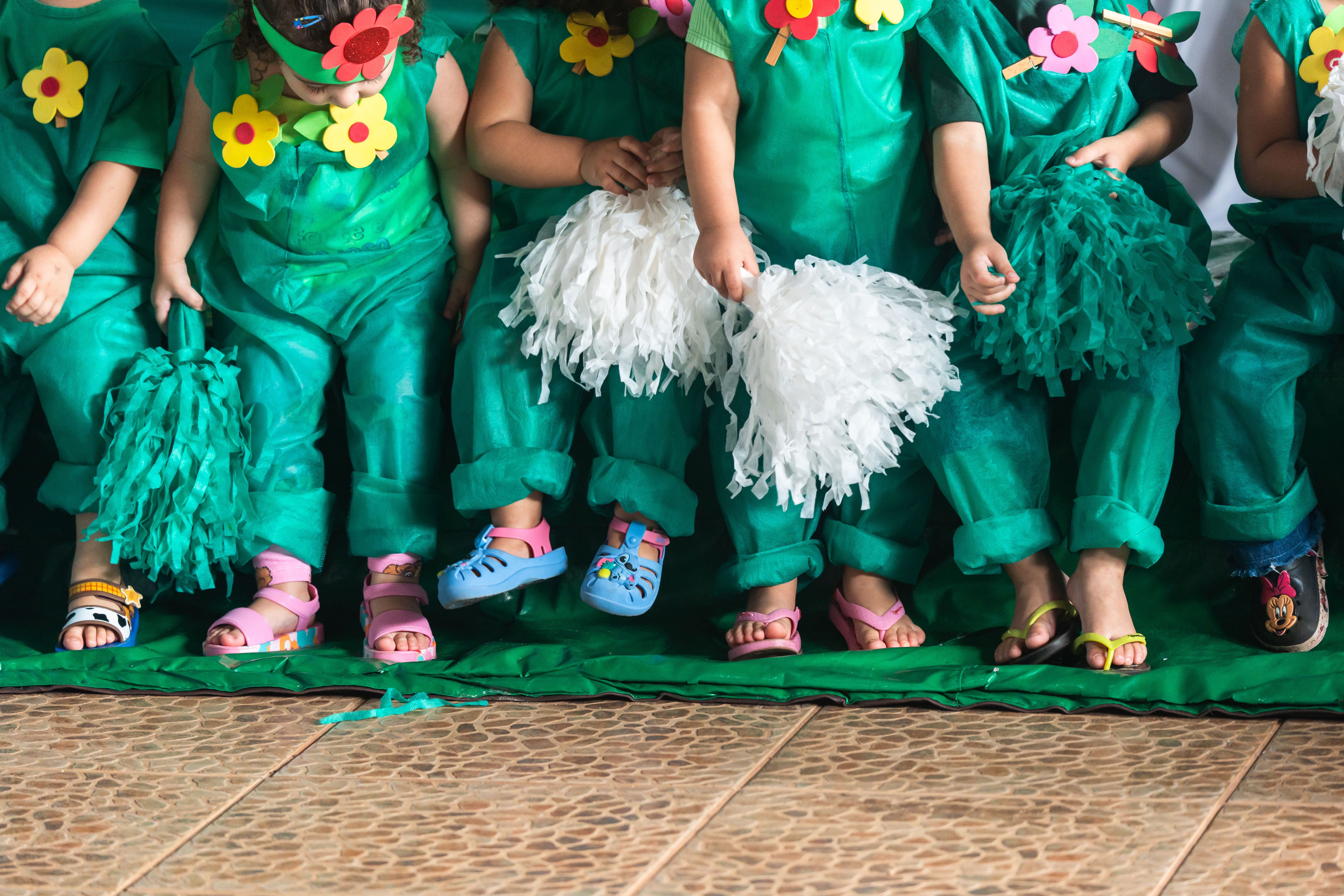Fotografia em plano médio cortada na altura do peito, mostrando uma fileira de crianças em pé, lado a lado. Elas vestem fantasias artesanais feitas de TNT verde, decoradas com flores coloridas de EVA coladas ou presas com grampos de roupa. Algumas crianças seguram pompons de papel nas cores branco e verde. O foco principal está nas pernas e nos pés das crianças, que usam sandálias coloridas de diversos estilos e cores, como rosa, azul e amarelo. Elas estão sobre um tapete verde estendido em um chão de pedras pequenas e rústicas. A imagem transmite a ludicidade de uma festa infantil.