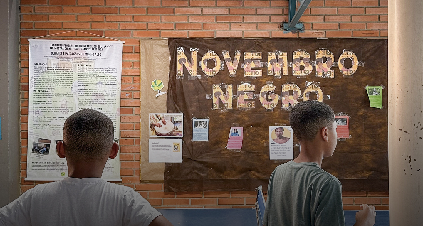 Fotografia em plano médio, de costas, mostra dois jovens estudantes negros observando painéis em uma parede de tijolos aparentes. À esquerda, há um banner científico do Instituto Federal do Rio Grande do Sul (IFRS). Ao centro, um grande painel de papel pardo exibe a frase "Novembro Negro" em letras recortadas e decoradas. Abaixo do título, estão coladas diversas fotos e textos informativos sobre personalidades e temas da cultura afro-brasileira. Os estudantes, um vestindo camiseta branca e o outro camiseta verde, contemplam o mural, que representa o trabalho pedagógico de valorização da ancestralidade no ambiente escolar.