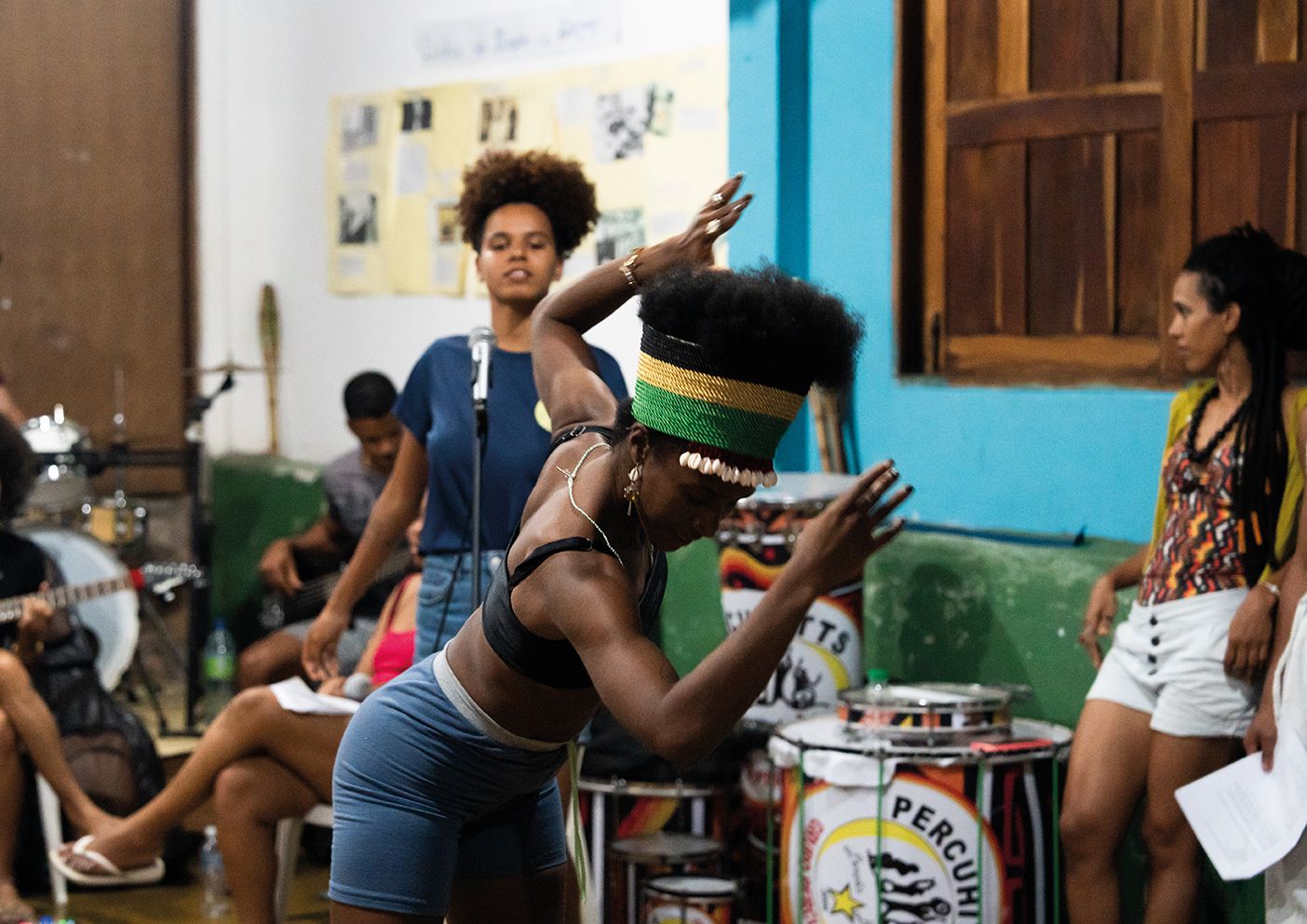 Fotografia em plano médio captura uma mulher negra dançando no centro de uma sala. Ela veste um top preto, bermuda azul e um turbante com as cores verde, amarelo e preto, decorado com búzios na testa. Seu corpo está inclinado para a frente com os braços erguidos em movimento. Ao fundo, outras pessoas acompanham a performance: uma mulher negra com cabelo afro fala ao microfone e, à direita, outra mulher observa o ensaio. Instrumentos musicais, como tambores de percussão com a inscrição