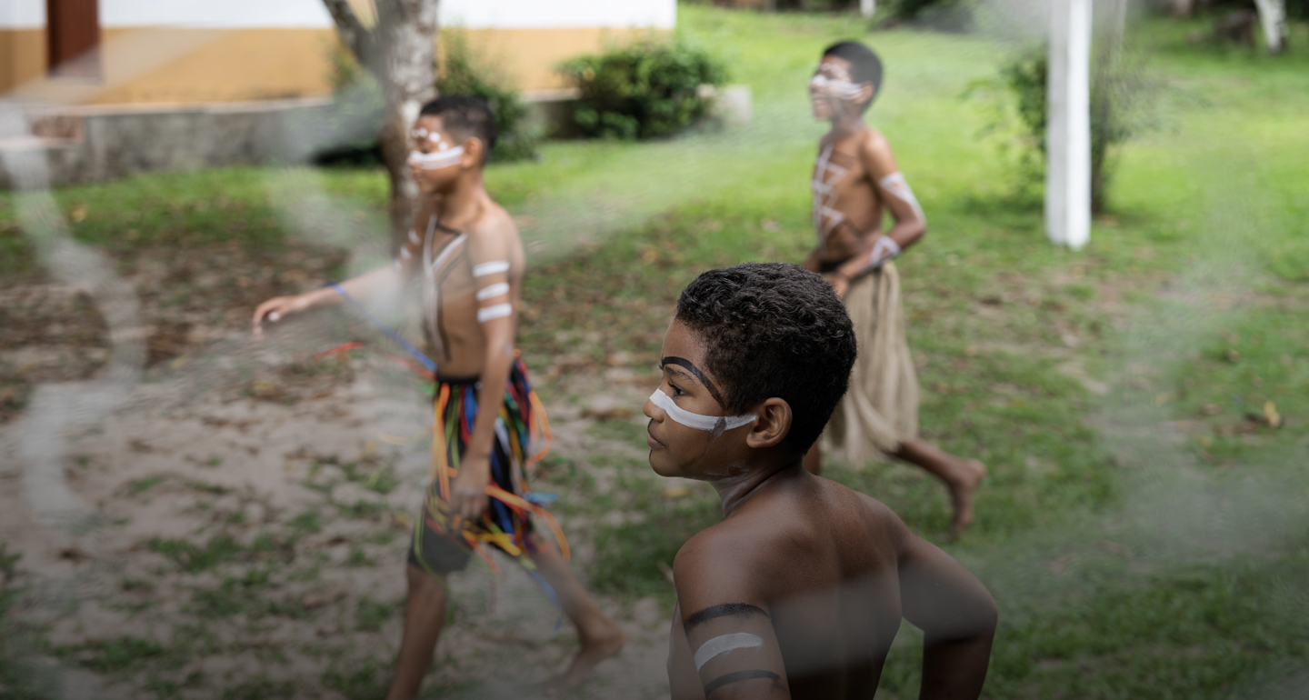 Foto em plano médio e profundidade de campo rasa, focada em um menino indígena em primeiro plano, de perfil, olhando para a esquerda. Ele tem pinturas tradicionais no rosto (linhas brancas e pretas) e no braço. Ao fundo, levemente desfocados, outros dois meninos com pinturas corporais e vestimentas típicas caminham e sorriem em um gramado. O cenário é externo, com árvores, grama e parte de uma construção simples ao fundo. Uma leve textura esfumaçada sobrepõe as bordas da imagem.