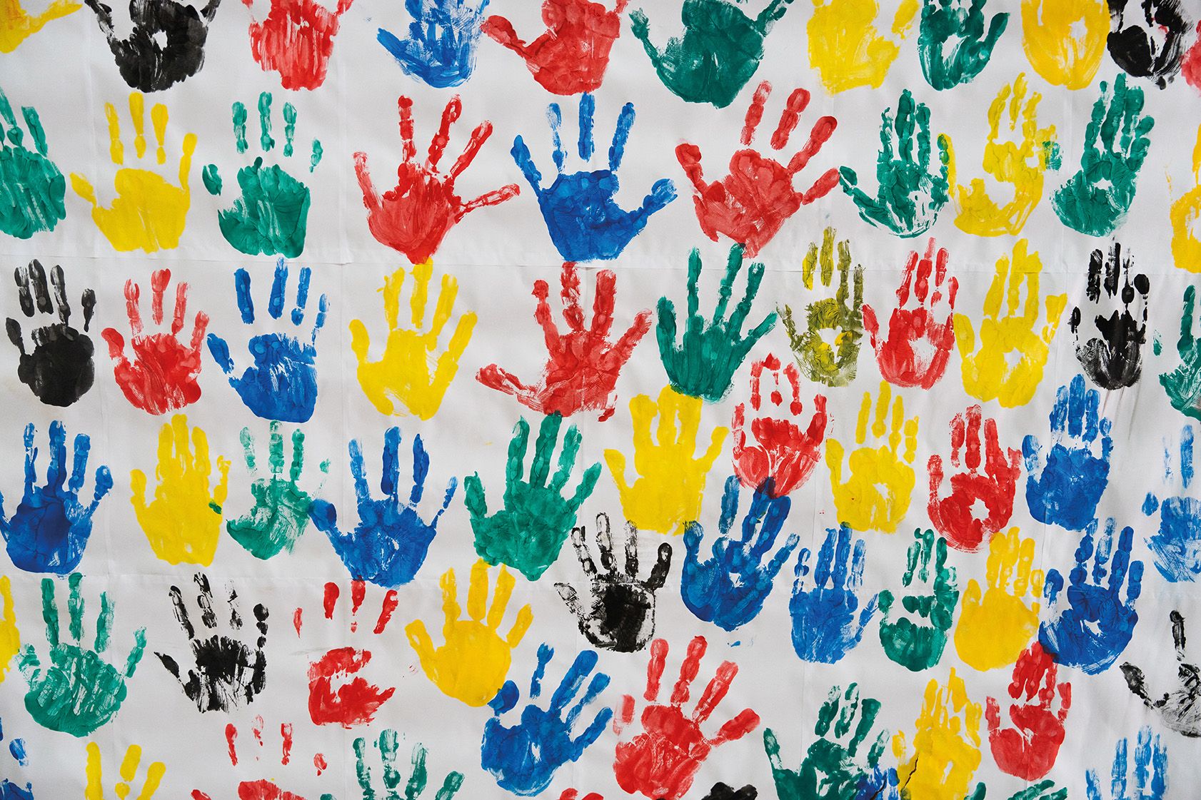 Foto em plano aberto de um grande painel de tecido branco estendido, preenchido por dezenas de impressões de palmas de mãos feitas com tinta guache. As marcas das mãos aparecem em diversas cores vibrantes como azul, amarelo, verde, vermelho e preto, espalhadas de forma irregular por toda a superfície. As impressões variam em tamanho e intensidade de tinta, criando um mosaico visual colorido e dinâmico que remete a uma atividade artística coletiva.