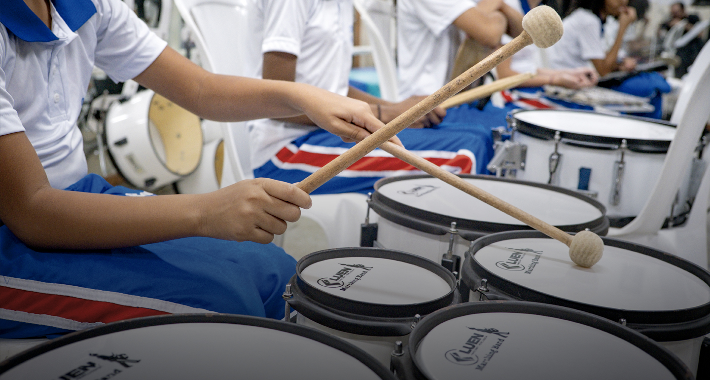 Foto em plano detalhe focada nas mãos de um estudante segurando baquetas de madeira cruzadas sobre tambores brancos de uma banda marcial. O jovem veste uniforme escolar com camiseta polo branca de gola azul e calça azul com listras laterais vermelhas e brancas. Ao fundo, outros estudantes sentados em cadeiras brancas aparecem desfocados, também com instrumentos de percussão. A imagem transmite ritmo e concentração. No rodapé, há uma leve sombra esfumaçada.