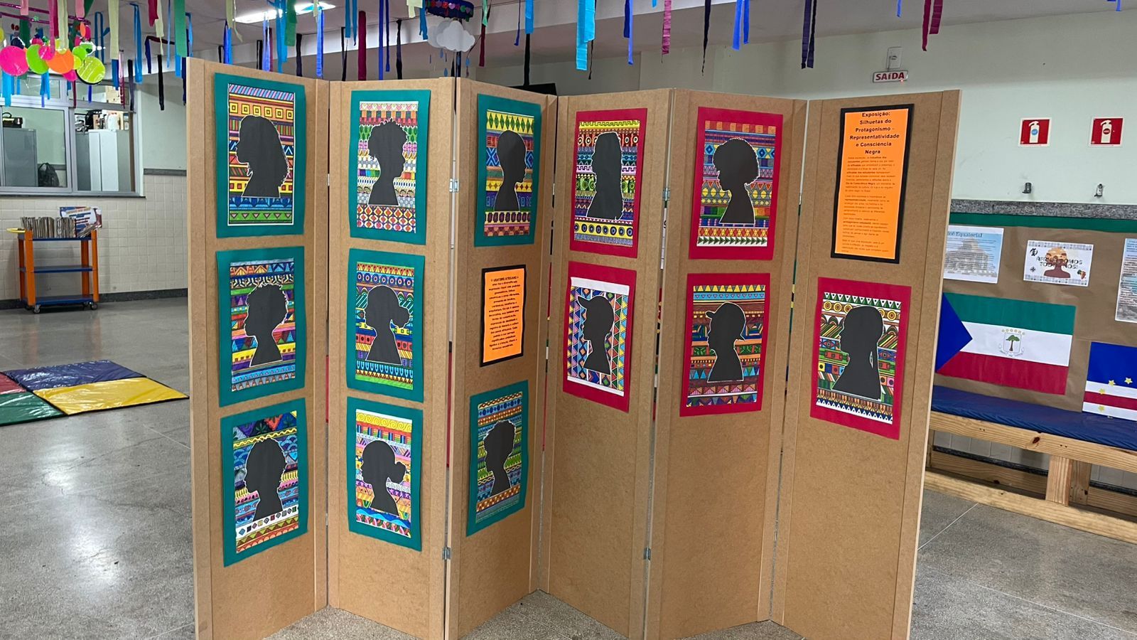 Foto de um painel expositor de madeira em formato de sanfona no centro de uma sala de aula ou saguão. O painel exibe diversos quadros com silhuetas negras de perfis humanos (homens, mulheres e crianças) em recortes de papel preto. Cada silhueta está centralizada sobre um fundo com padrões geométricos e tribais coloridos. Há também cartazes laranjas com textos explicativos. Ao fundo, o ambiente escolar é decorado com fitas coloridas penduradas no teto e bandeiras de países africanos na parede à direita. O chão é de granilite cinza claro.