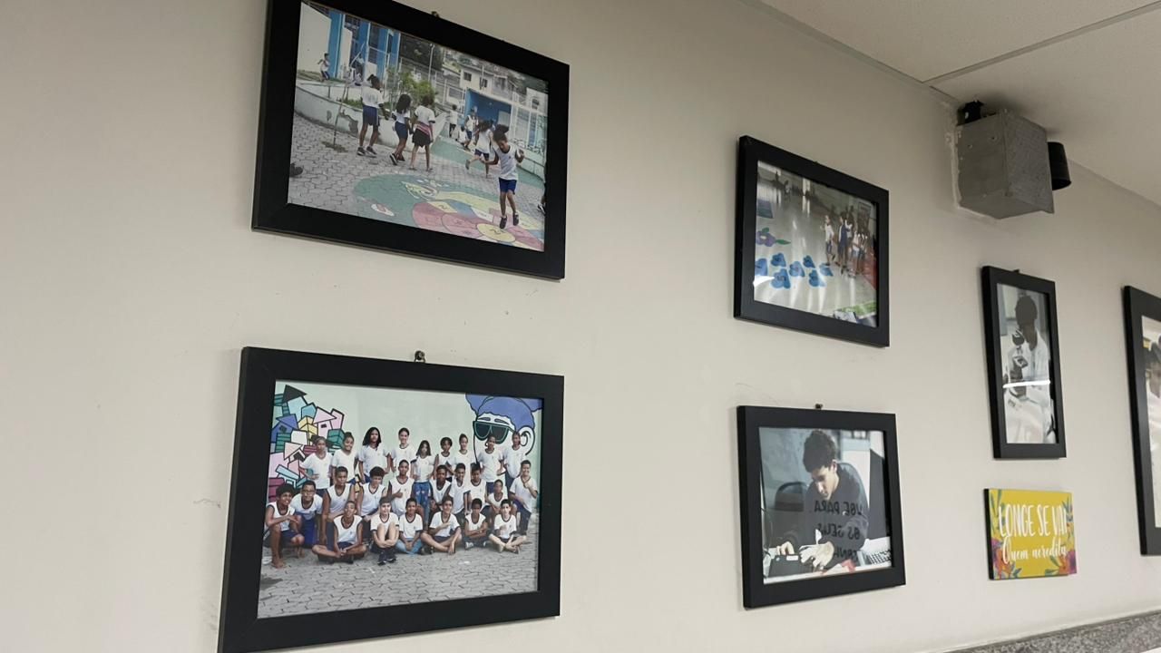 Foto em &acirc;ngulo diagonal de uma parede branca decorada com quadros de moldura preta. As fotografias nos quadros mostram momentos da vida escolar: crian&ccedil;as brincando em um p&aacute;tio externo colorido, um retrato em grupo de alunos uniformizados em frente a um grafite, e estudantes em atividades de sala de aula. &Agrave; direita, h&aacute; um quadro vertical com uma foto em preto e branco e uma pequena placa colorida com uma frase motivacional. A ilumina&ccedil;&atilde;o &eacute; suave e vem do teto, criando uma galeria afetiva no corredor da institui&ccedil;&atilde;o.