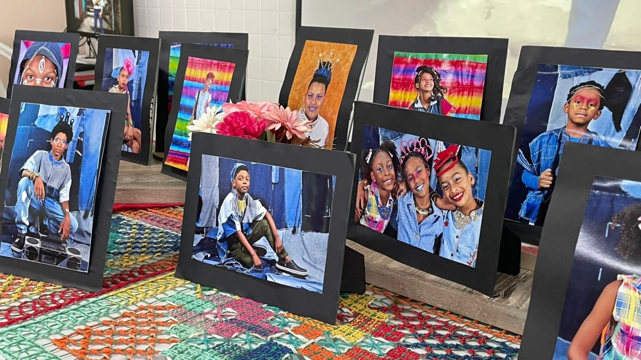 Foto em plano médio de uma exposição de arte. Diversos retratos fotográficos com molduras de papel preto estão organizados sobre uma superfície forrada com um tapete de crochê colorido em tons de amarelo, vermelho e verde. As fotos mostram crianças e jovens negros em poses imponentes, vestindo roupas jeans, acessórios prateados, coroas e maquiagens faciais artísticas. Ao centro, um pequeno arranjo de flores artificiais em tons de rosa e branco decora a mesa. O fundo está levemente desfocado, sugerindo um ambiente escolar ou cultural.