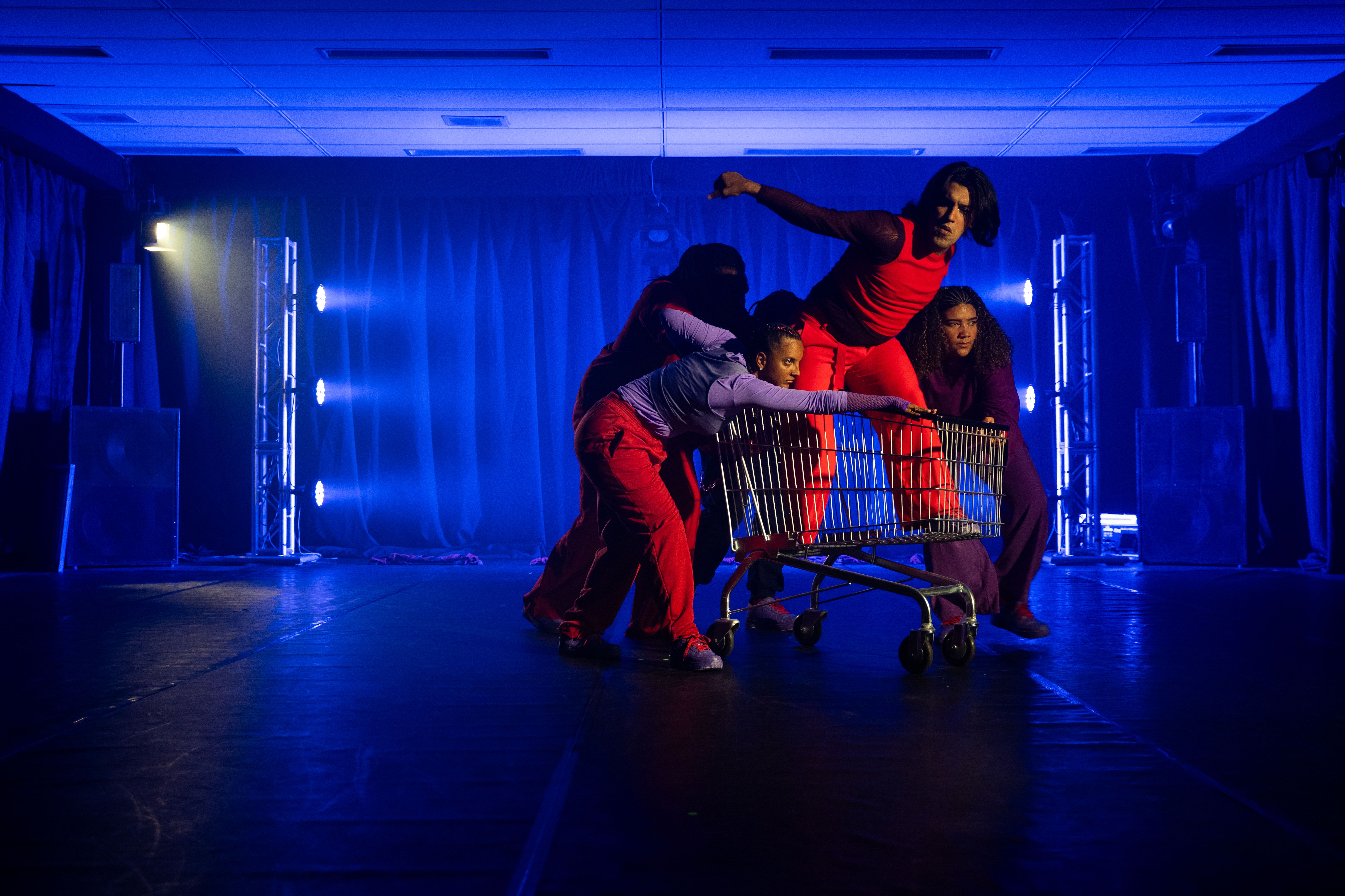 Uma cena de performance artística em um palco sob forte iluminação azul. No centro, quatro pessoas interagem com um carrinho de compras metálico. Um homem de vermelho está em pé dentro do carrinho, com o tronco inclinado para frente. Ao redor, três artistas em roupas vibrantes (roxo, lilás e vermelho) empurram ou se apoiam no objeto com expressões intensas.

O cenário tem cortinas ao fundo e torres de luz laterais que emitem feixes azuis horizontais. O chão é escuro e reflexivo, criando uma atmosfera dramática e contemporânea de movimento e tensão coletiva.