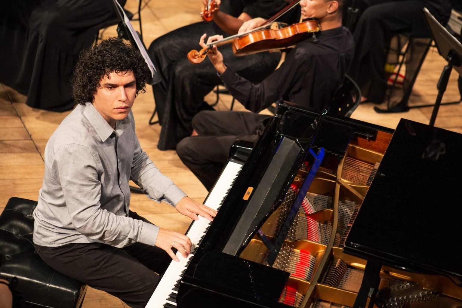 A imagem mostra um pianista em primeiro plano durante um concerto. Ele &eacute; um homem jovem, de pele clara e cabelos pretos cacheados, vestindo uma camisa social cinza. Ele est&aacute; sentado em frente a um piano de cauda preto aberto, com as m&atilde;os sobre as teclas brancas. Ao fundo, m&uacute;sicos de uma orquestra aparecem parcialmente, tocando instrumentos de corda como o violino. O ambiente tem ilumina&ccedil;&atilde;o quente, focada no pianista, e o ch&atilde;o &eacute; de madeira clara. A cena transmite concentra&ccedil;&atilde;o e seriedade