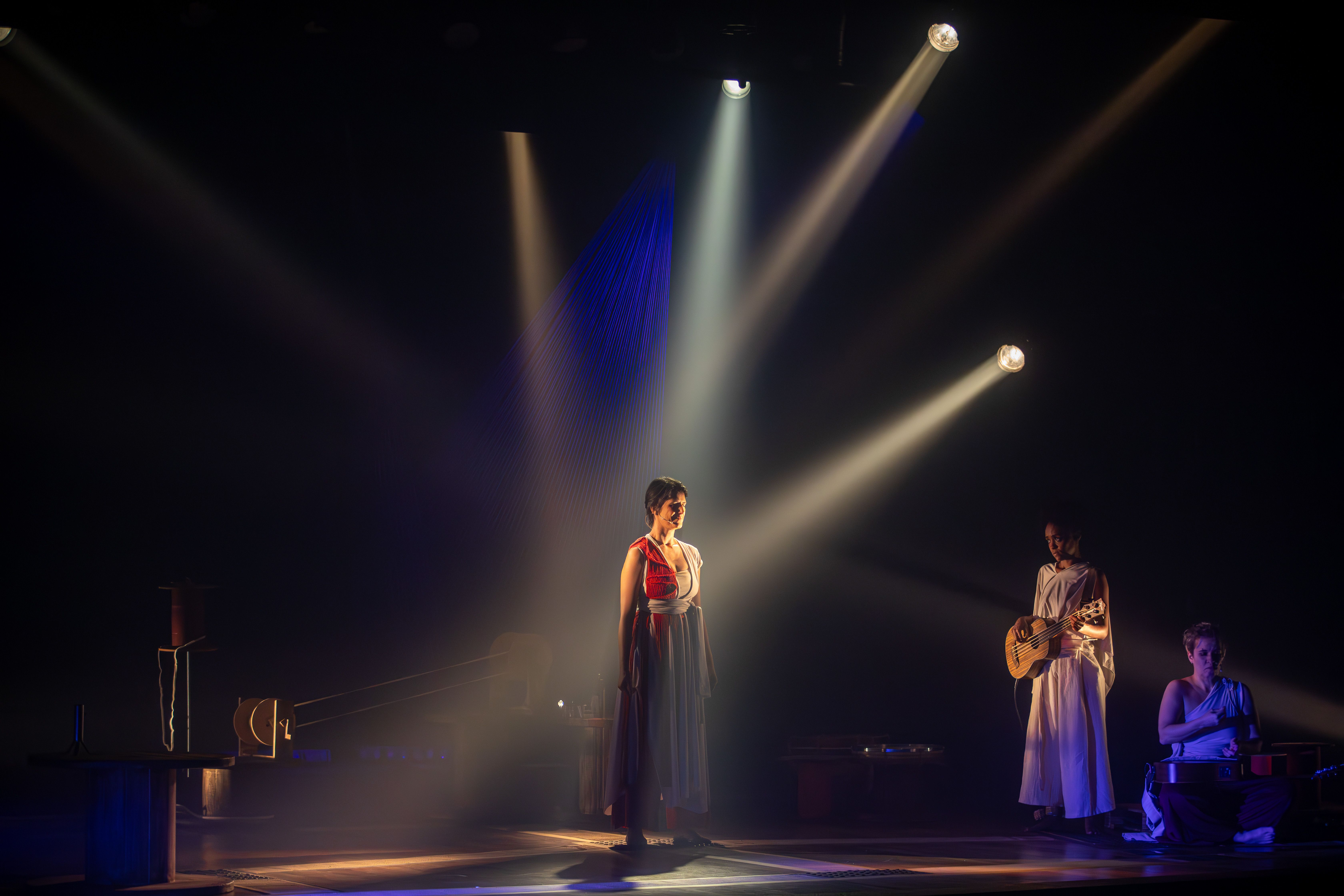 
Em um palco escuro, tr&ecirc;s mulheres realizam uma performance sob feixes de luz direcionados. Ao centro, uma mulher de pele clara veste um longo vestido branco com detalhes em vermelho, iluminada por um refletor superior. &Agrave; direita, uma mulher negra em p&eacute; veste trajes brancos e toca um instrumento de cordas, enquanto outra mulher est&aacute; sentada ao ch&atilde;o, operando equipamentos de som. Feixes de luz branca cruzam o cen&aacute;rio, e ao fundo projeta-se um cone de linhas azuis vibrantes. O clima &eacute; solene e teatral