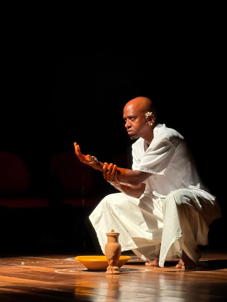 Uma fotografia em plano m&eacute;dio, com fundo preto, mostra um homem negro, calvo e descal&ccedil;o, agachado em um palco de madeira. Ele veste uma roupa branca folgada e adornos na orelha direita. Seus bra&ccedil;os e m&atilde;os est&atilde;o manchados de um l&iacute;quido marrom-alaranjado, enquanto ele olha fixamente para a palma da m&atilde;o direita elevada. No ch&atilde;o, &agrave; sua frente, h&aacute; uma tigela dourada ao lado de uma pequena moringa de cer&acirc;mica sobre um c&iacute;rculo desenhado com p&oacute; branco. A ilumina&ccedil;&atilde;o focal destaca o performer e os objetos ritual&iacute;sticos